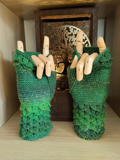 A pair of green crocheted fingerless gloves displayed on wooden mannequin hands. The gloves feature crocodile stitch detailing at the wrist in a gradient of green shades, transitioning from dark to light. The palm section is worked in a smooth, flat stitch, while the top near the fingers returns to the textured crocodile stitch. The gloves have a scalloped edge at the wrist. The mannequin hands are posed with pinky and index fingers raised in a “rock on” gesture. A decorative wooden box with a metallic tree design sits in the background, adding an artistic touch to the display.