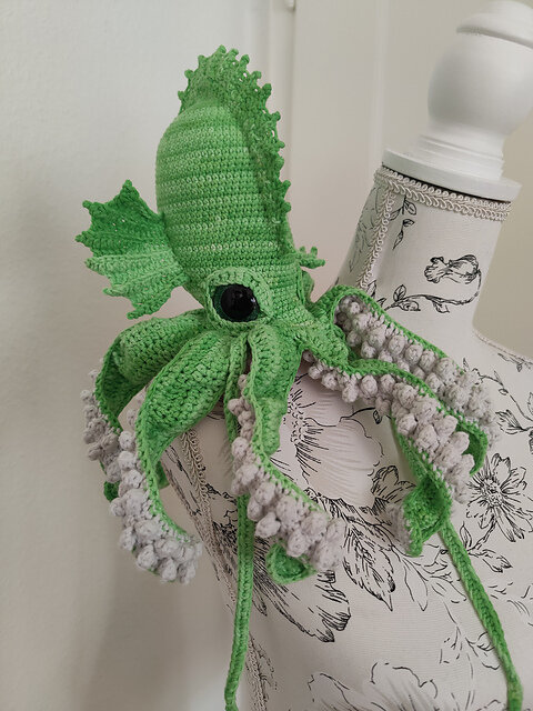 A crocheted sea creature resembling a squid or kraken is draped over a white mannequin torso printed with black floral designs. The creature is made from light green yarn with long, curling tentacles whose undersides feature rows of small, textured light gray suction cups. It has one large, glossy black safety eye on the side of its head and two small crocheted fins positioned behind the eye. The body and tentacles curve naturally over the mannequin, showing the shaping and stitch texture of the crochet work. The background is softly lit and out of focus.