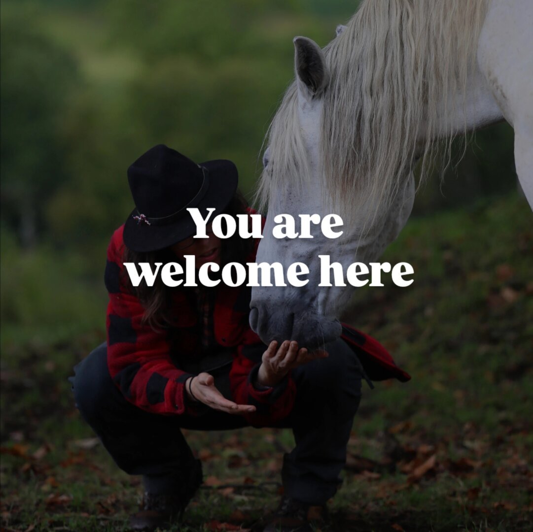 A woman with a hat and chequered jacket is squatting on a green field. Next to her there is a horse. The horse gently nuzzles her hand.