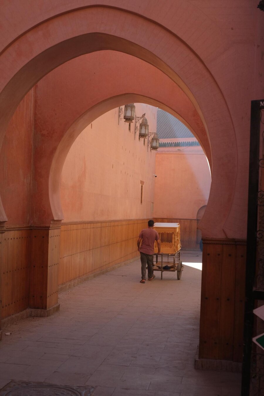 Un homme pousse une charette de dos, sous les voûtes rose pales de la médina.