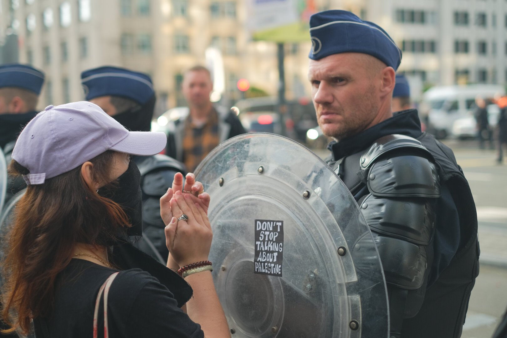 Lors d'une manifestation devant l'ambassade des États-Unis de Bruxelles. Un policier avec un grand bouclier rond et transparent. Le policier a les sourcils froncés et une moue concentrée et pas content. sur son bouclier, il y a un sticker, un rectangle noir, écrit en blanc 