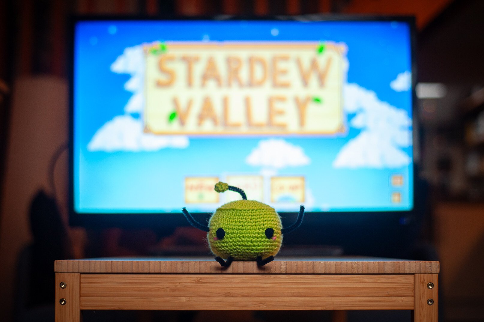 A green crochet junimo from stardew valley in front of a big screen with the stardew valley menu page on it