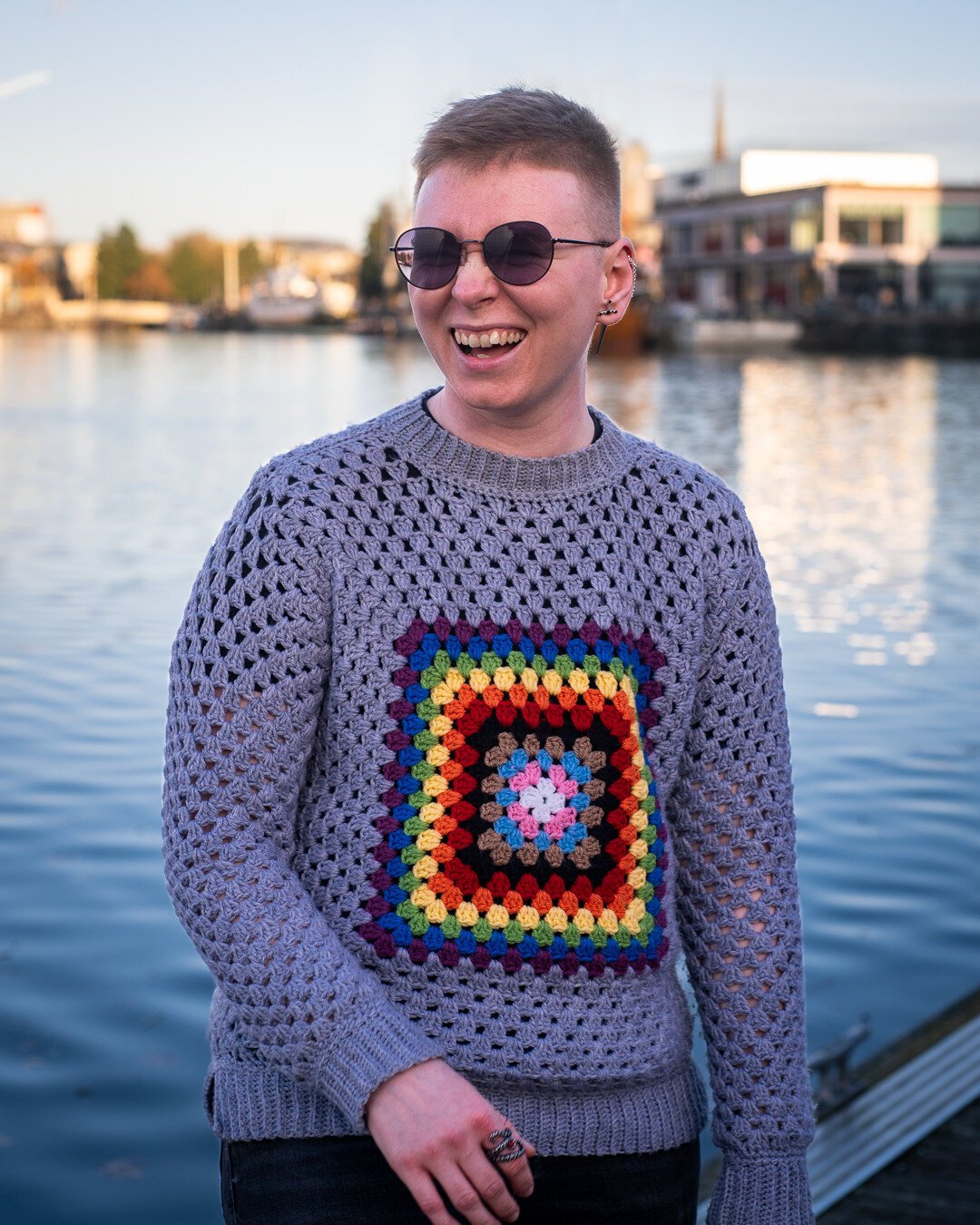 Nicky stands in front of a harbour front on a sunny day, wearing sunglasses and a handmade grey crochet jumper with a progress Pride flag in the middle. They are laughing and looking off camera