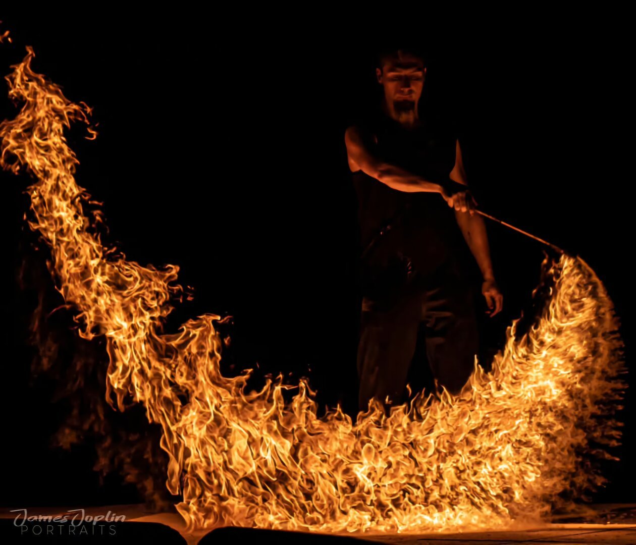 "A fire performer dressed in black, creating a dramatic arc of flames in the darkness with a fire staff."