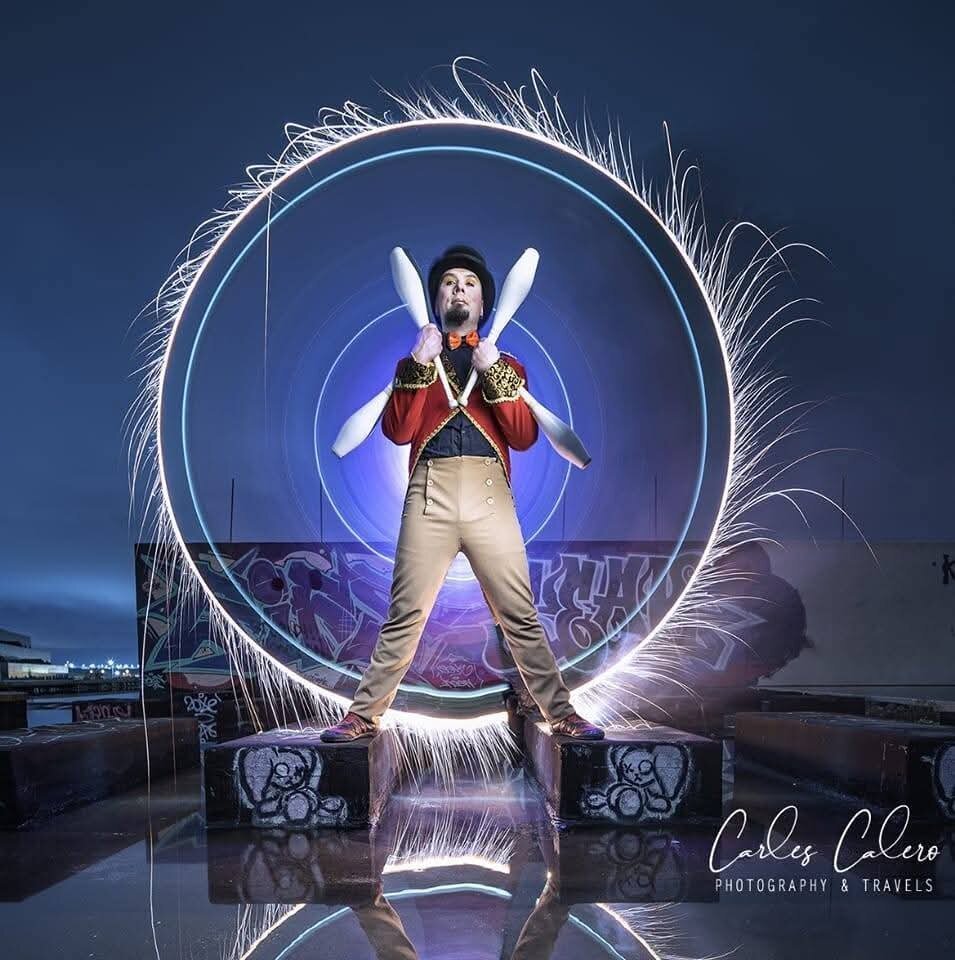 A circus artist stands confidently on a graffiti-covered rooftop at dusk, dressed in a red and gold ringmaster outfit with a black top hat and orange bow tie. Holding four white juggling clubs crossed over his chest, he looks directly at the camera. Behind him, a mesmerizing light painting effect creates a glowing circular halo with blue and white neon-like rings, while sparks fly outward, adding a dynamic energy to the scene. The wet rooftop reflects the light, enhancing the dramatic atmosphere. In the bottom right corner, the photographer's watermark reads "Carles Calero Photography & Travels."