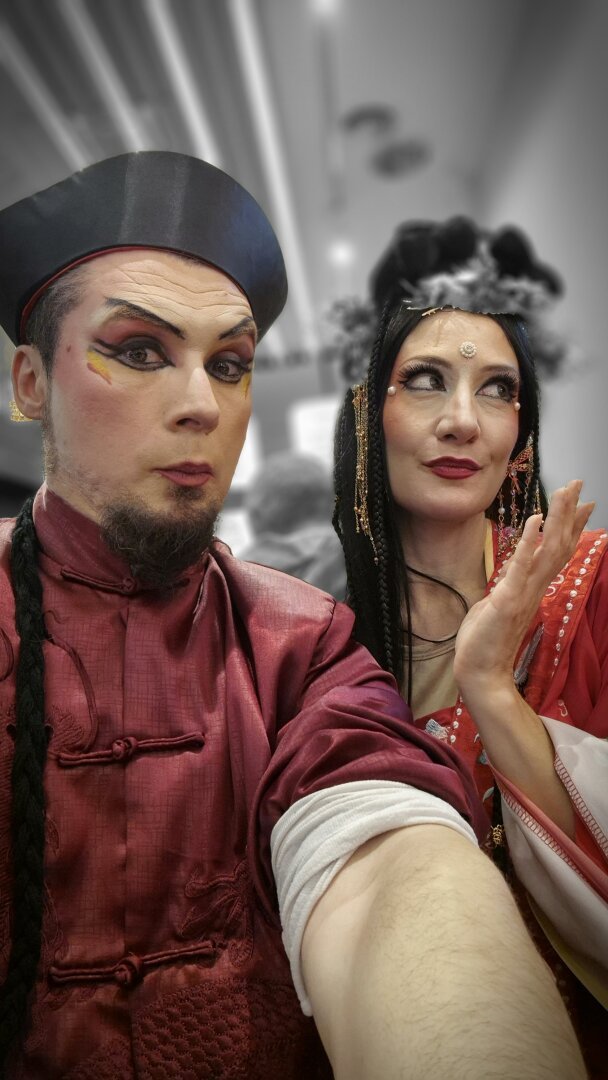 A festive selfie of two circus artists dressed in elaborate Chinese-inspired costumes for the Chinese New Year parade in Usera, Madrid. The man on the left wears a deep red silk outfit with intricate embroidery, a traditional black hat, bold theatrical makeup, and a long braided hairstyle. The woman on the right