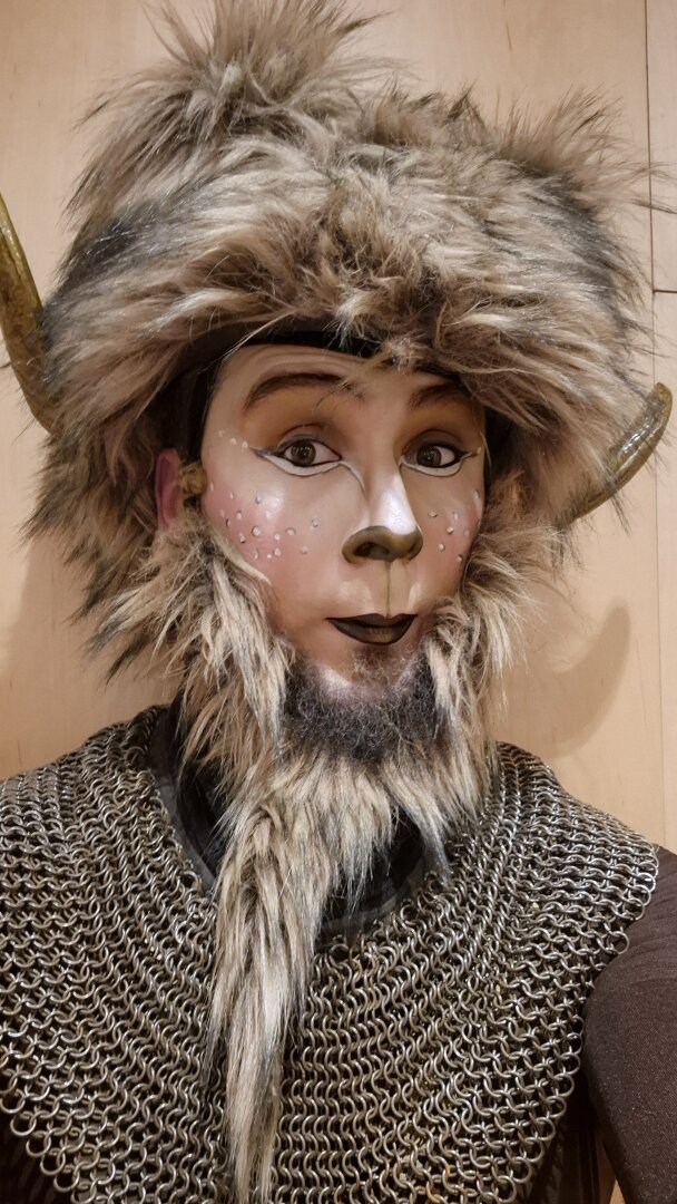 A close-up portrait of a performer in a fantasy-inspired costume resembling a mythical creature. The face is painted with artistic makeup, featuring white accents around the eyes, small decorative dots on the cheeks, and a dark-painted nose and lips. The performer wears a furry headpiece with small horns and a thick faux-fur beard. The costume includes a chainmail shirt, adding a medieval or fantasy warrior aesthetic. The performer's expression is playful and expressive, with wide eyes and a slightly surprised look."