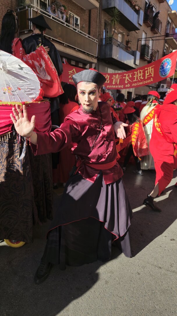 A circus artist in a striking red and black traditional outfit performs in the Chinese New Year parade in Usera, Madrid. He strikes a dynamic martial arts-inspired pose with one hand extended, captivating the crowd with his intense expression and theatrical makeup. Behind him, a vibrant parade of red costumes, banners with Chinese characters, and performers with fans and umbrellas bring the celebration to life. Spectators watch from balconies, adding to the festive atmosphere. 🐉🎭🔥