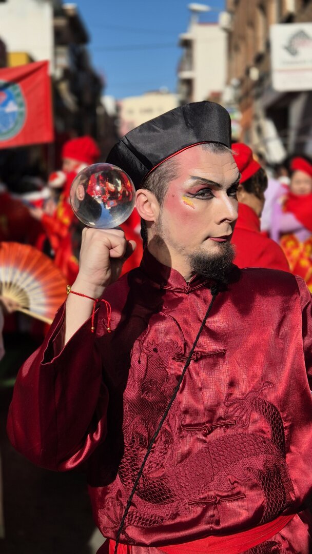 A circus artist in an elegant red silk outfit with intricate dragon embroidery performs contact juggling during the Chinese New Year parade in Usera, Madrid. He holds a reflective crystal ball in one hand, creating a mesmerizing illusion as the surrounding crowd and red decorations appear mirrored inside it. His intense gaze and theatrical makeup add to the mystique of the performance. Behind him, parade participants dressed in red with traditional fans and banners fill the streets, celebrating the Year of the Snake. 🐉✨🎭🔥