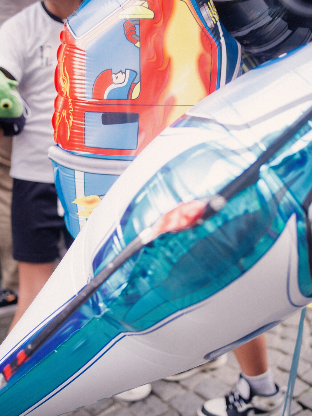 A street photograph of glossy inflatable foil balloons at an outdoor festival. A white and cyan rocket balloon creates a sharp diagonal line across the foreground, leading toward a bright red and yellow toy fire truck balloon behind it. In the background, pedestrians walk across a paved cobblestone street in natural daylight.