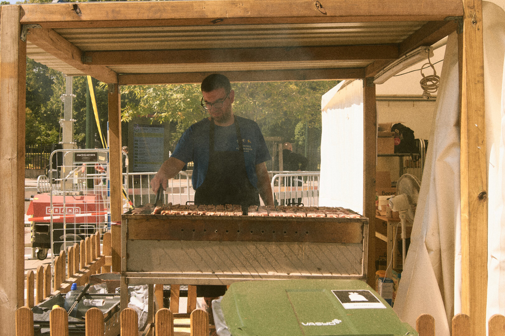A man standing under a wooden frame, cooking numerous meat skewers over a large charcoal grill.
