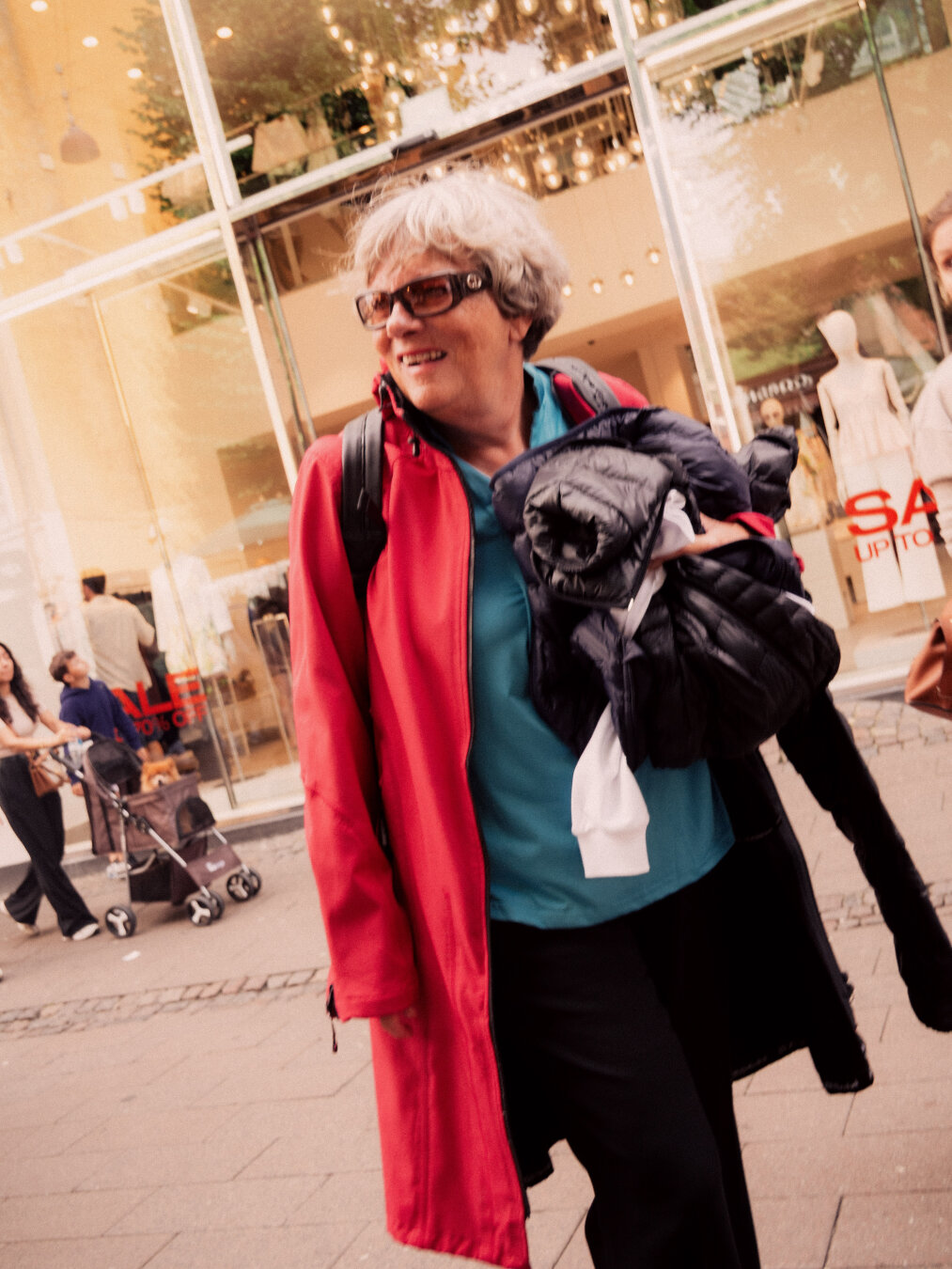 A candid, slightly tilted portrait of a grey-haired woman smiling in a bright red coat over a blue shirt as she walks past a sunlit storefront displaying a "SALE" sign.
