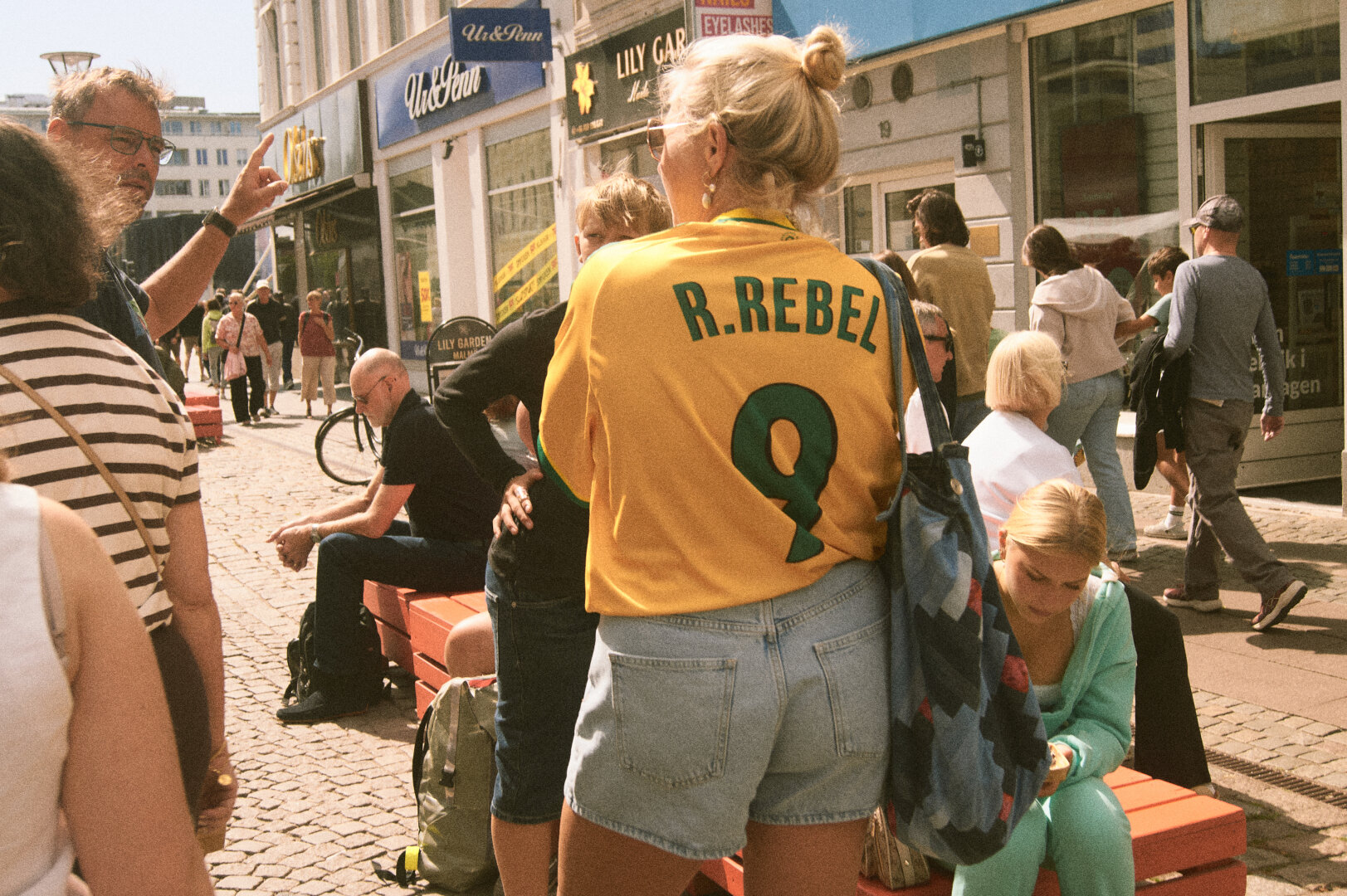 A crowded, sunny pedestrian street scene featuring a woman in the foreground wearing a bright yellow jersey with "R.REBEL 9" printed on the back.