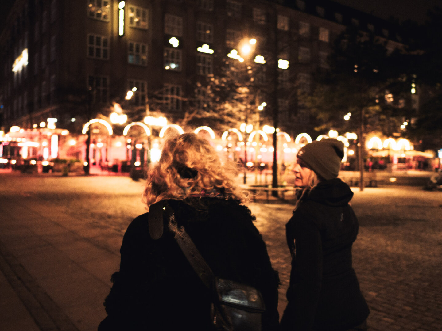 Night street photography. From behind, two women walk through a cobbled city square illuminated by warm, golden, out-of-focus café lights. The image has a cinematic, grainy, and slightly blurry atmospheric quality.
