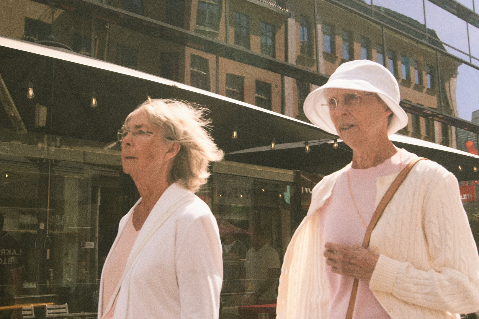 Two older women walking through a sunlit city street, displaying a warm, grainy, film-like aesthetic.