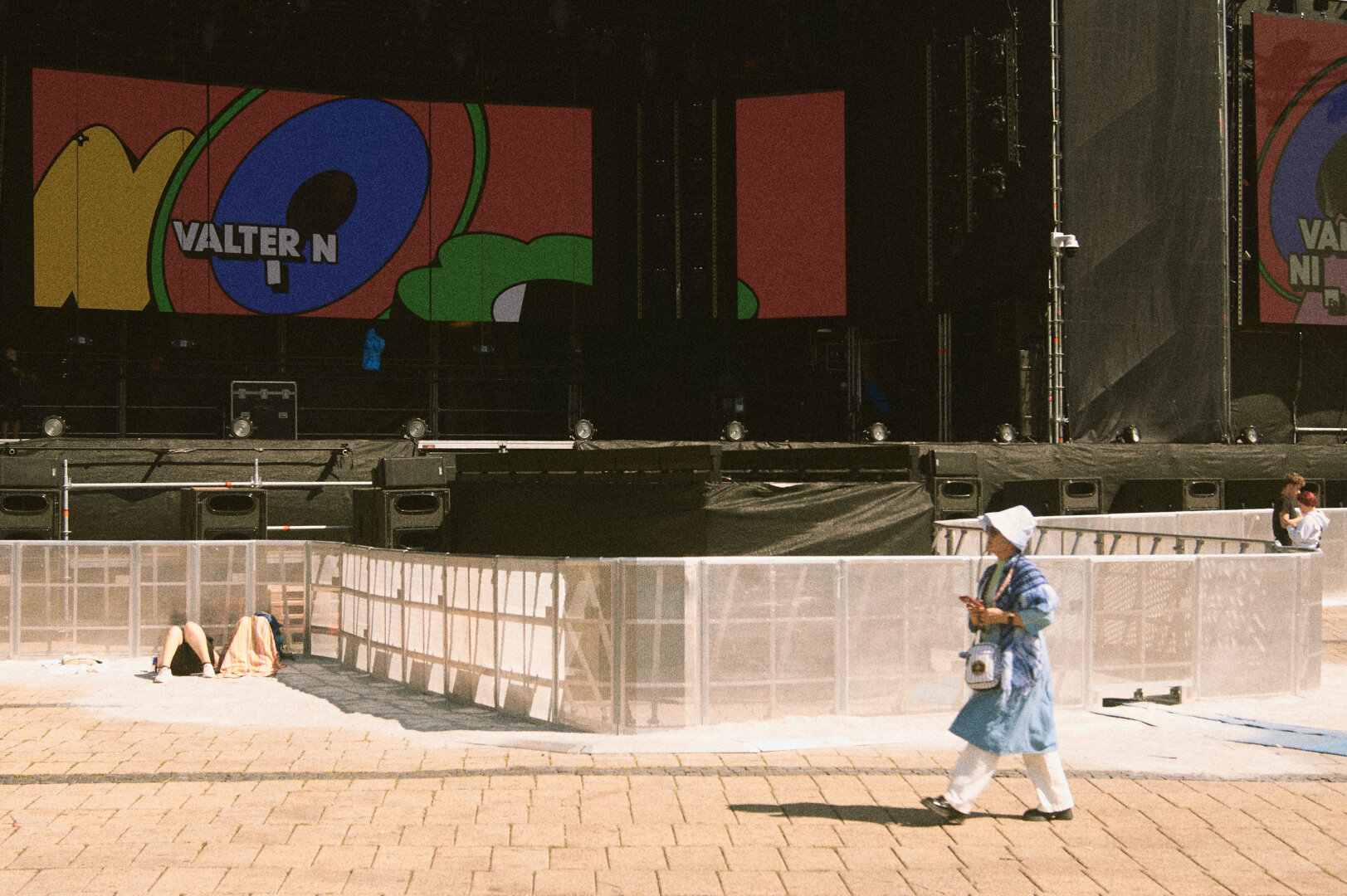 A person wearing a blue coat and sun hat walking past metal barriers in front of a large, colorful festival stage screen.