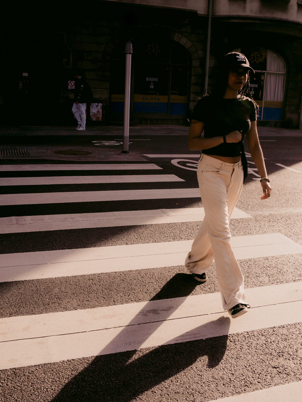 A high-contrast street photograph taken in bright, directional sunlight showing a woman wearing an Adidas cap and wide-leg white pants walking across a zebra crosswalk in Malmö. Her body is caught mid-stride, casting a long, dark diagonal shadow onto the striped road surface behind her. The background store facades sit in deep shadow.