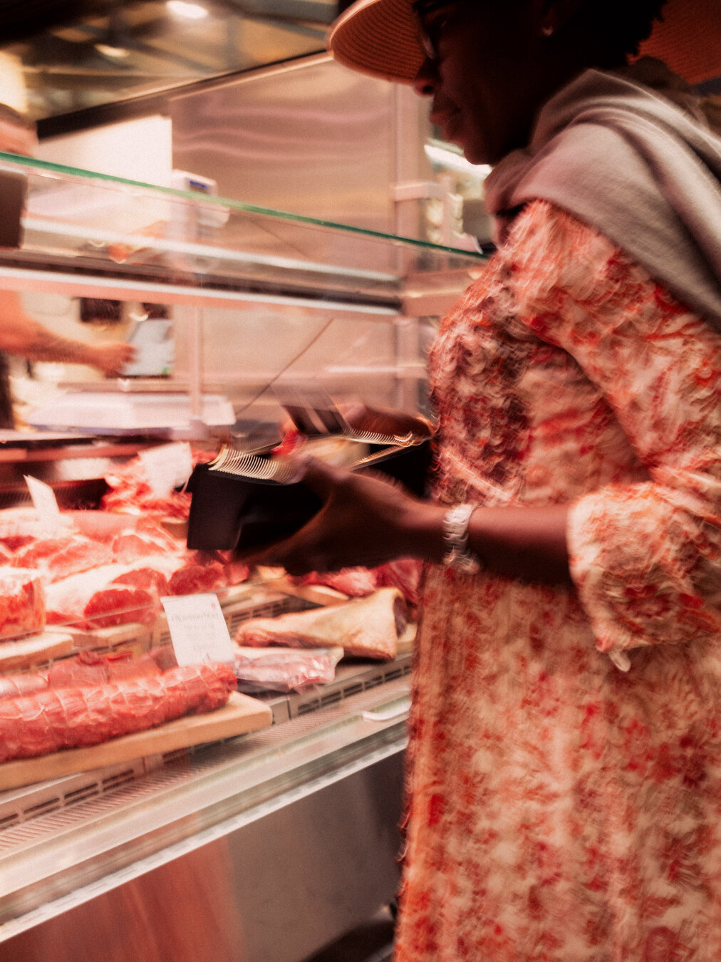 A close-up side profile of a woman wearing a red-patterned dress and straw hat at a glass butcher display case, reaching toward raw red meats with natural motion blur.