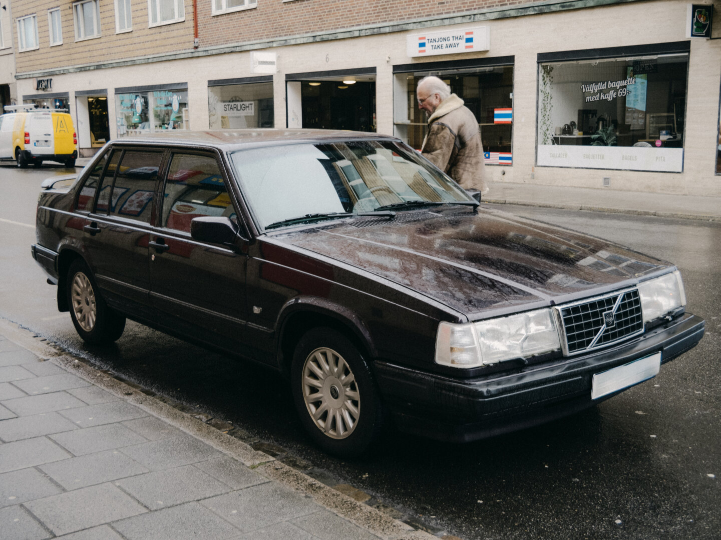 A dark vintage Volvo sedan parked on a wet asphalt street in Malmö, Sweden. Behind the car, a man in a beige shearling coat walks past storefronts under an overcast sky.