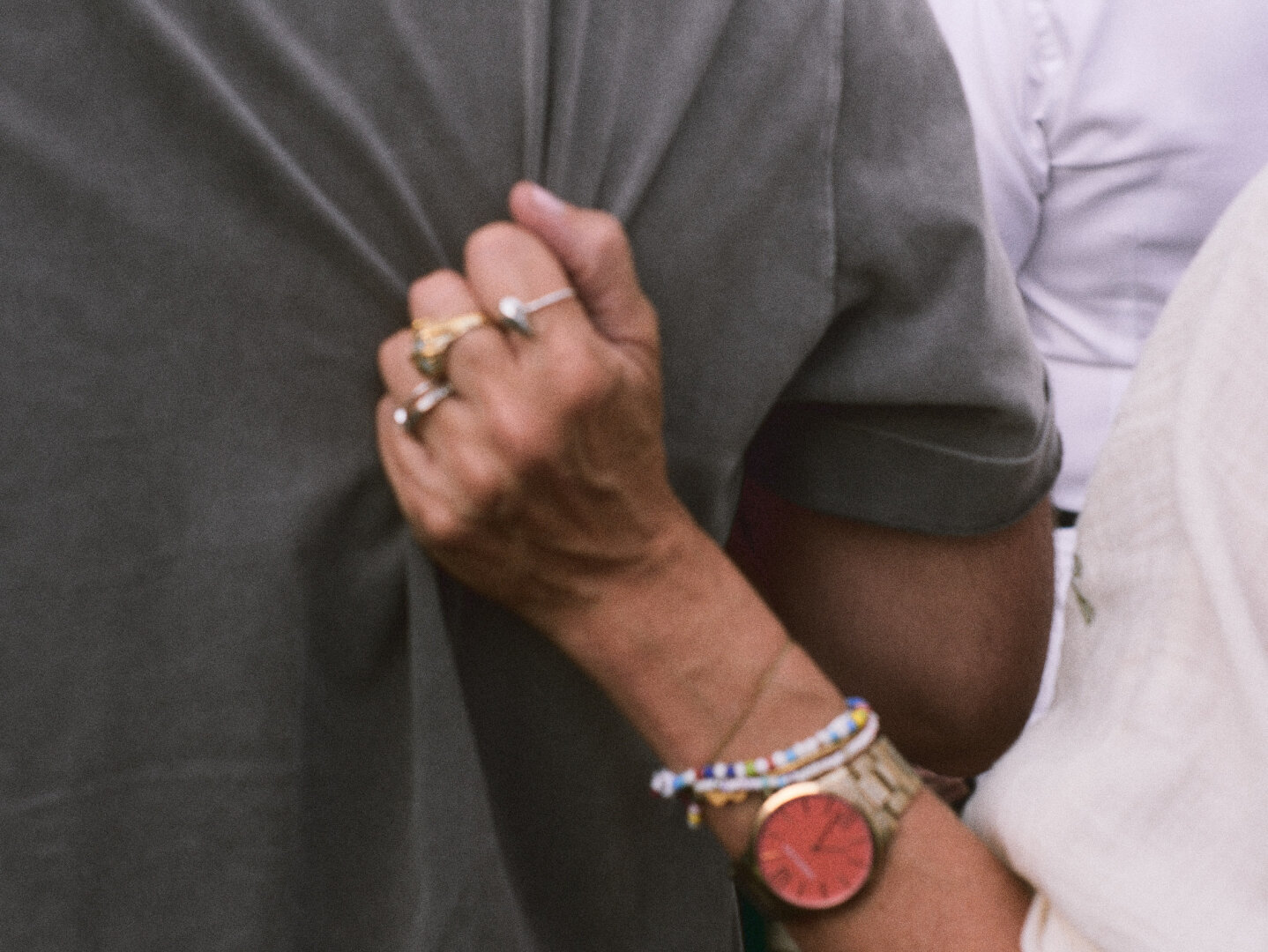 Close-up street photograph of a hand tightly gripping the back of someone's grey shirt. The hand belongs to someone wearing multiple rings, colorful beaded bracelets, and a gold watch with a bright red dial. The framing is tightly cropped, capturing the tension of the gesture and the texture of the fabric.