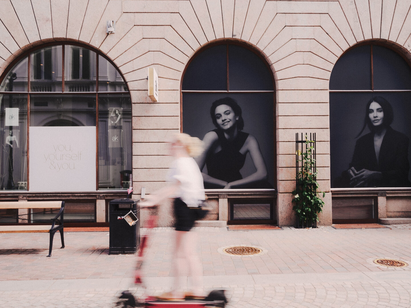 A street photography image showing a heavily blurred person riding a red electric scooter from right to left. The background is sharp, featuring a stone building with three large arched windows. The windows display a sign reading "you, yourself & you." next to two large, high-fashion black and white portraits of women. The slow shutter speed creates a contrast between the fast-moving rider and the static storefront.