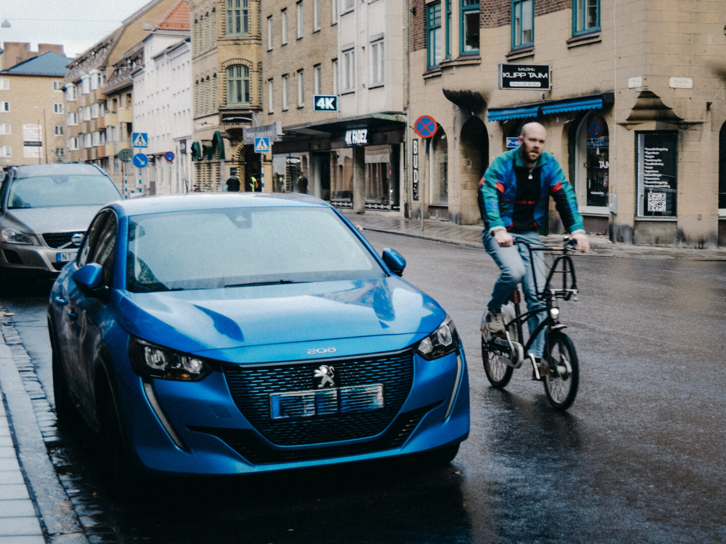 A vibrant electric blue Peugeot hatchback parked on a rainy city street in Malmö, Sweden, as a cyclist in a matching blue and red jacket rides past in motion.