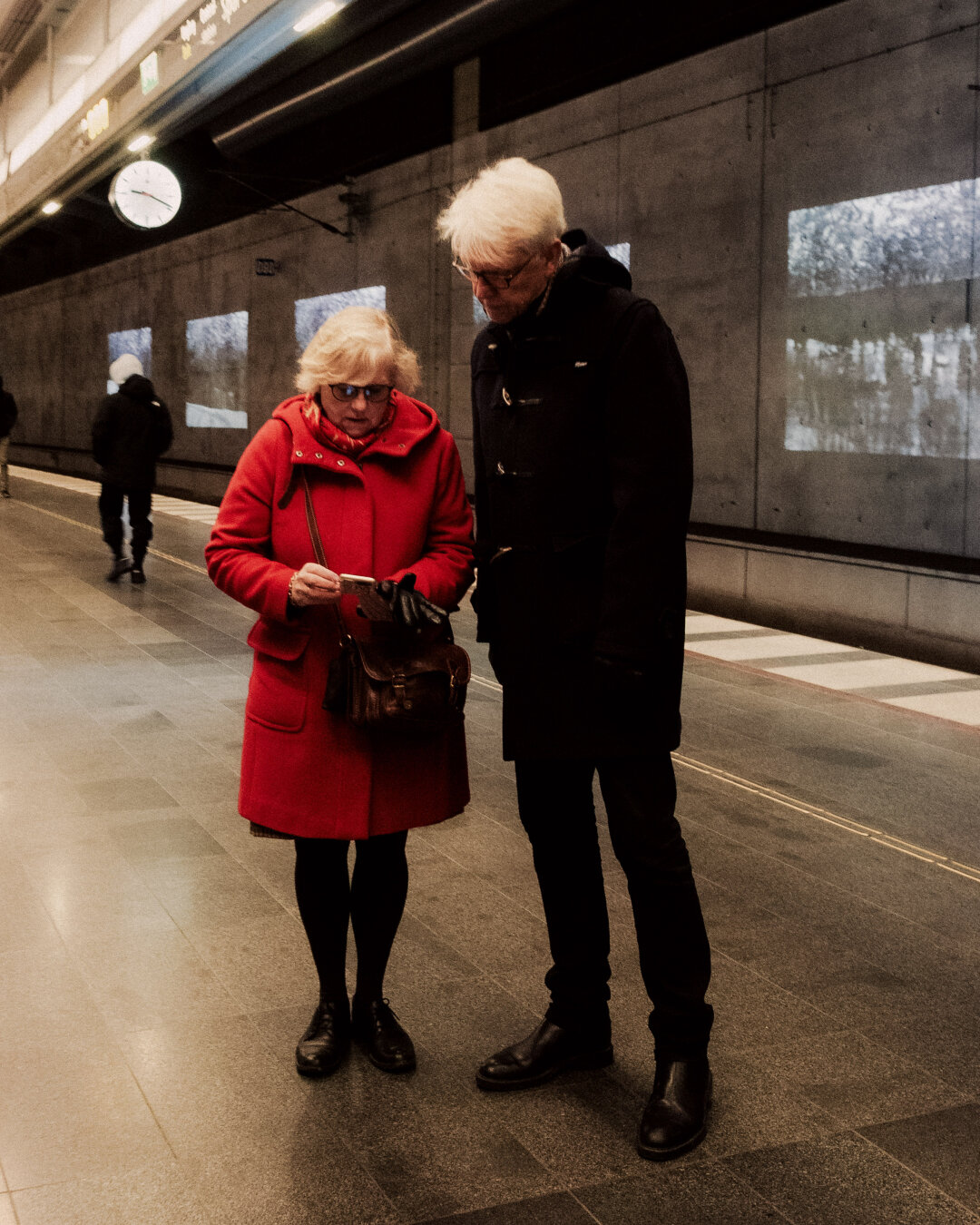 An older couple standing on a concrete underground train platform; the woman wears a vibrant bright red winter coat while examining a phone, contrasted against cold grey walls.