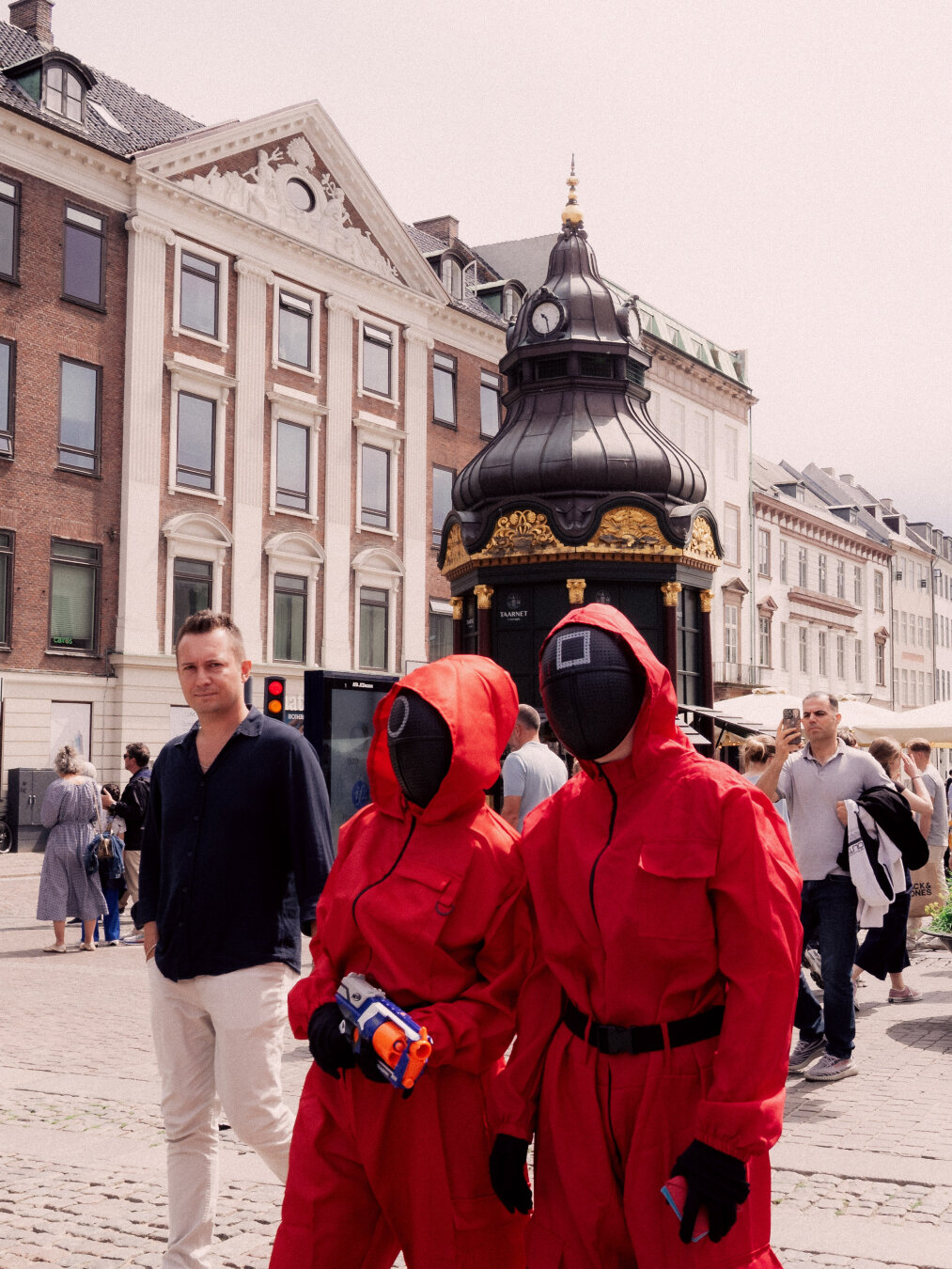 Two street performers dressed in bright red Squid Game jumpsuits and black geometric masks holding a toy gun on a cobbled square, with historical Danish architecture in the background.