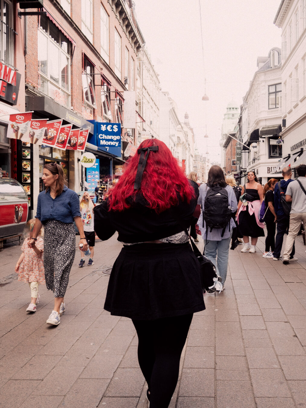 Back-view of a person walking down a bustling European pedestrian street, featuring long, intensely dyed red hair with a black bow in a high-contrast street scene.