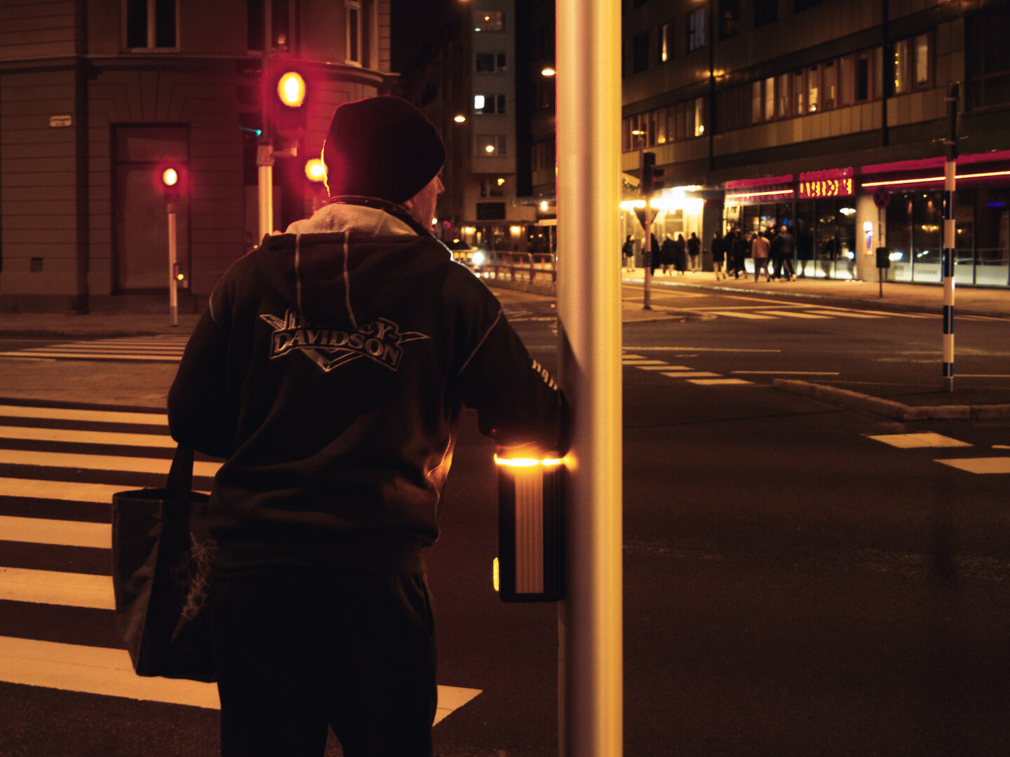 Night street photography. A person wearing a black beanie and a Harley Davidson hoodie stands at a crosswalk at night, shot from behind. They are resting their hand on a glowing yellow crosswalk button. A bright red traffic light shines in the background against dark city buildings.