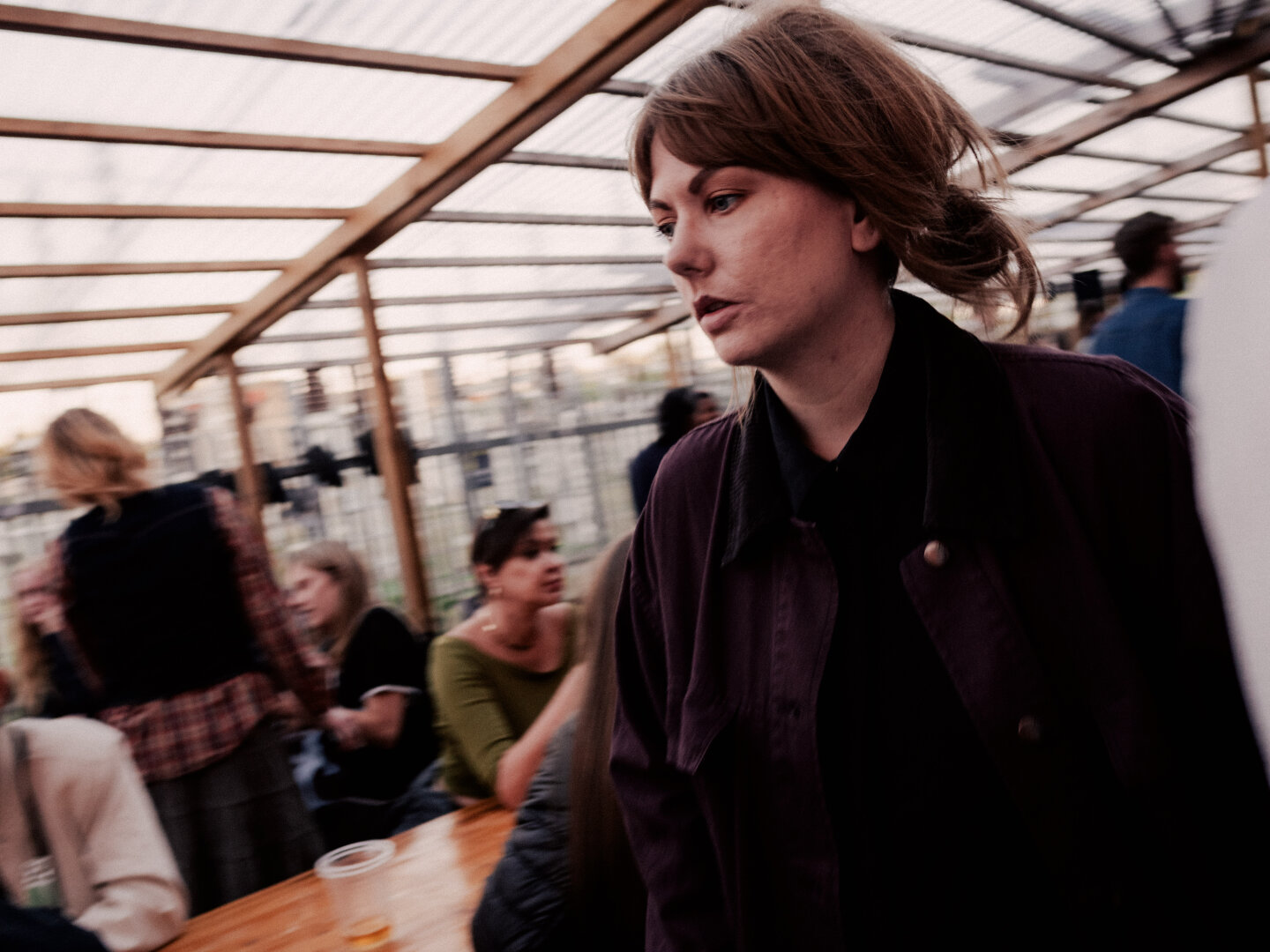 A candid street photograph of a woman with light brown hair wearing a dark purple shirt, walking outdoors underneath a wooden pergola structure. Other festival attendees sit in the blurred background.