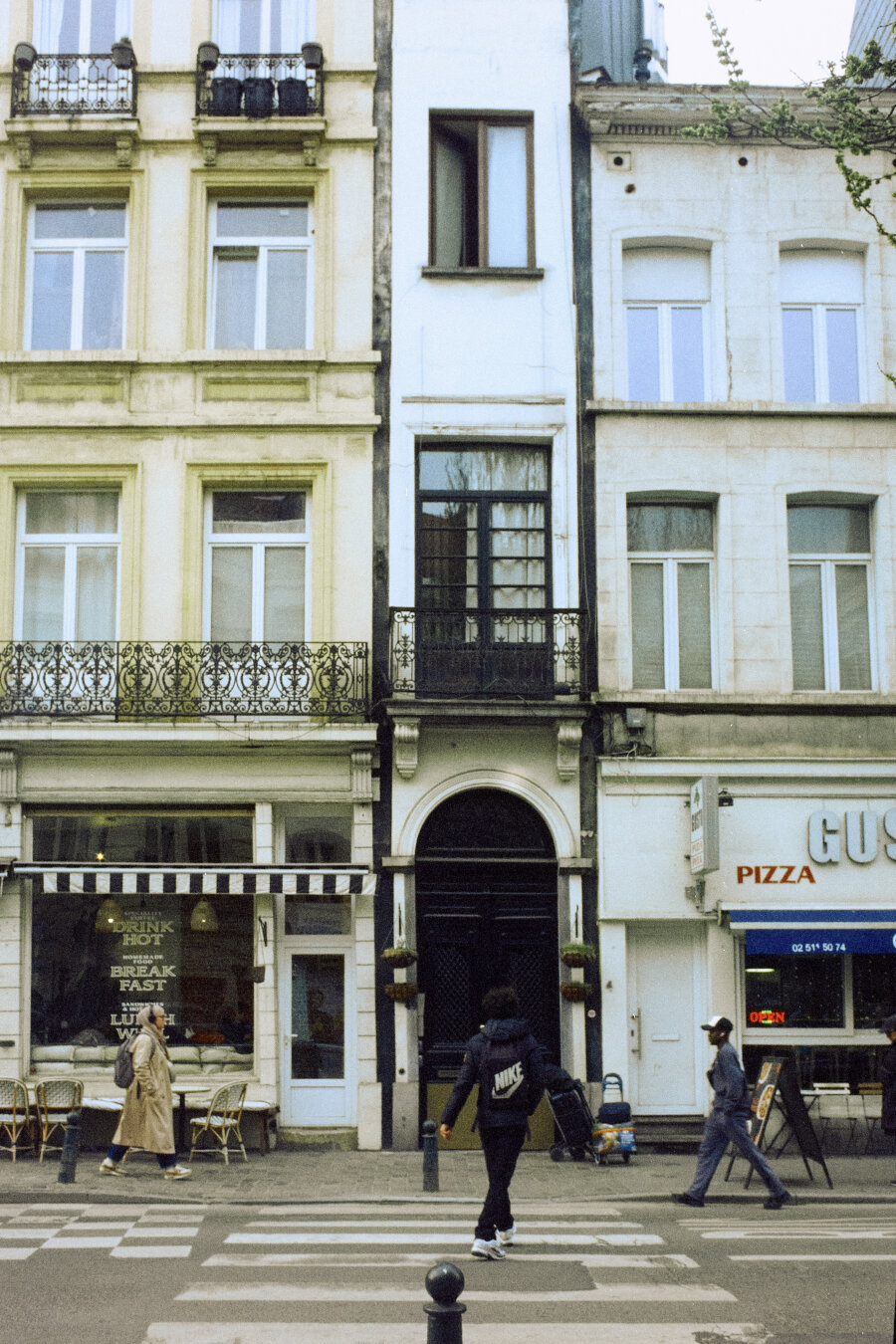 Analog street photography. A perfectly centered, impossibly thin white building is sandwiched between two wider, classical European facades in Brussels. In the foreground, three pedestrians cross a zebra crossing in different directions. The colors have a soft, cinematic film aesthetic.