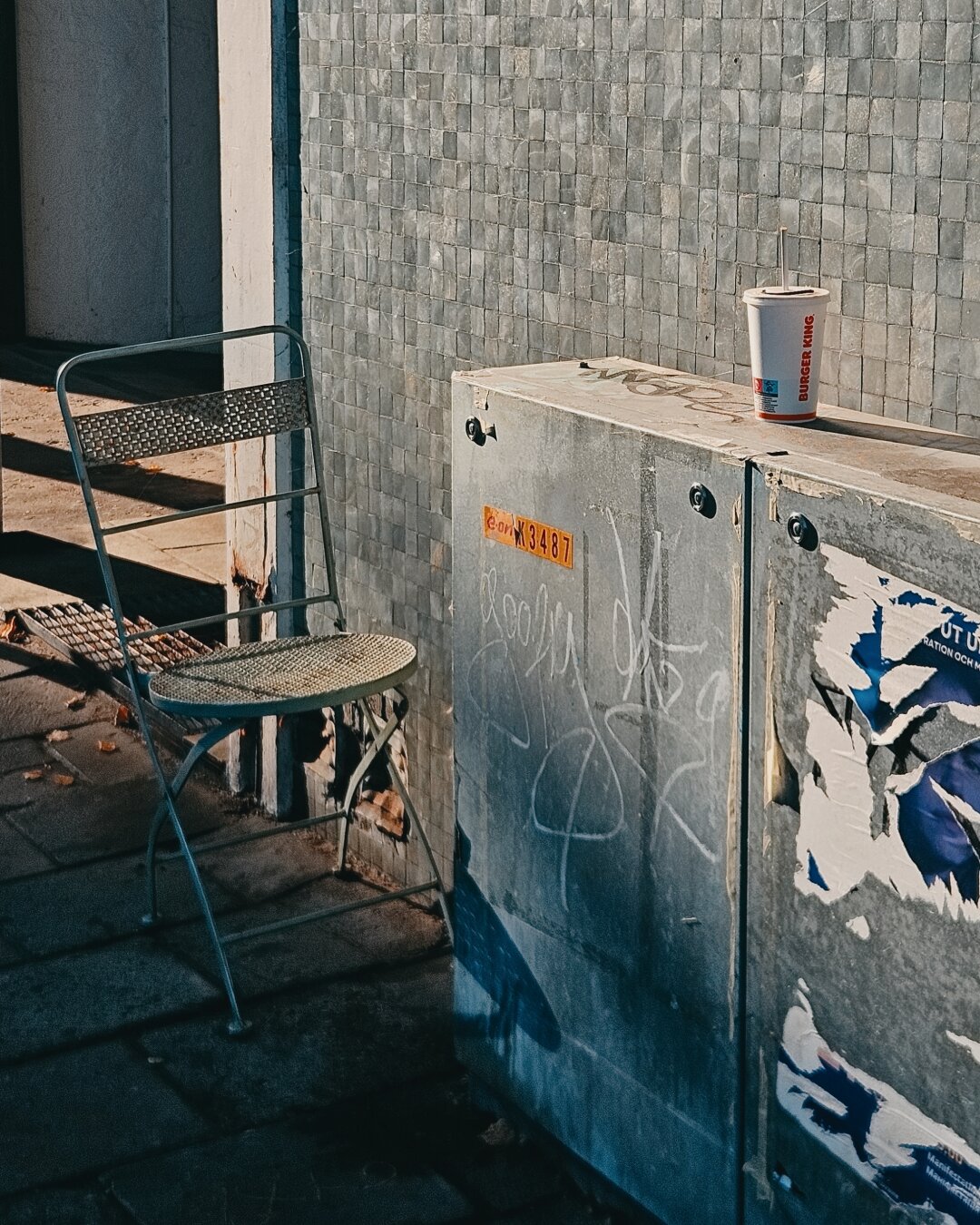 High-contrast street photograph taken in Malmö under bright afternoon sunlight. A light-colored metal folding chair sits in shadow next to an grey utility box covered in torn posters and graffiti tags. A disposable paper soda cup sits on top of the utility box, catching warm sunlight against a textured grey wall.