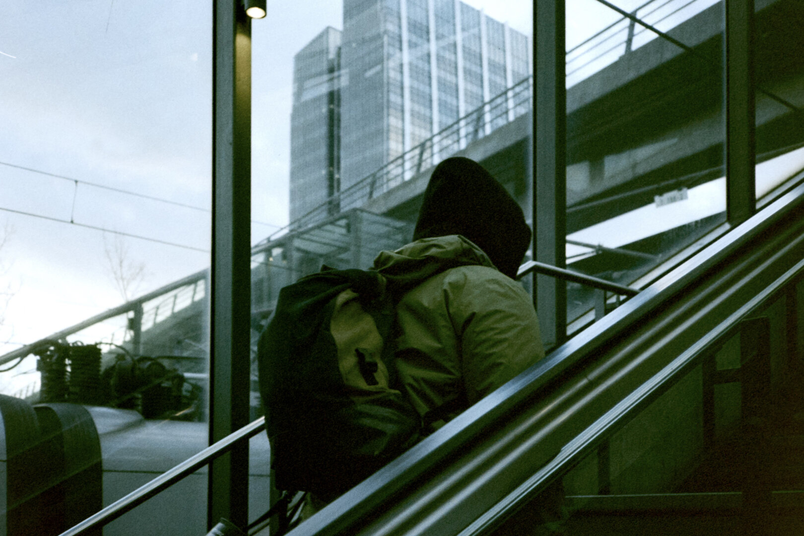 A grainy color film photograph with a distinct green cast showing an anonymous person in an olive green hooded jacket and backpack riding an escalator up inside a glass-enclosed train station. The composition relies on the strong diagonal lines of the escalator rails set against the perfectly leveled horizontal and vertical lines of a concrete overpass and a modern glass building outside. A white emulsion scratch and scan dust are intentionally left visible in the top left sky.