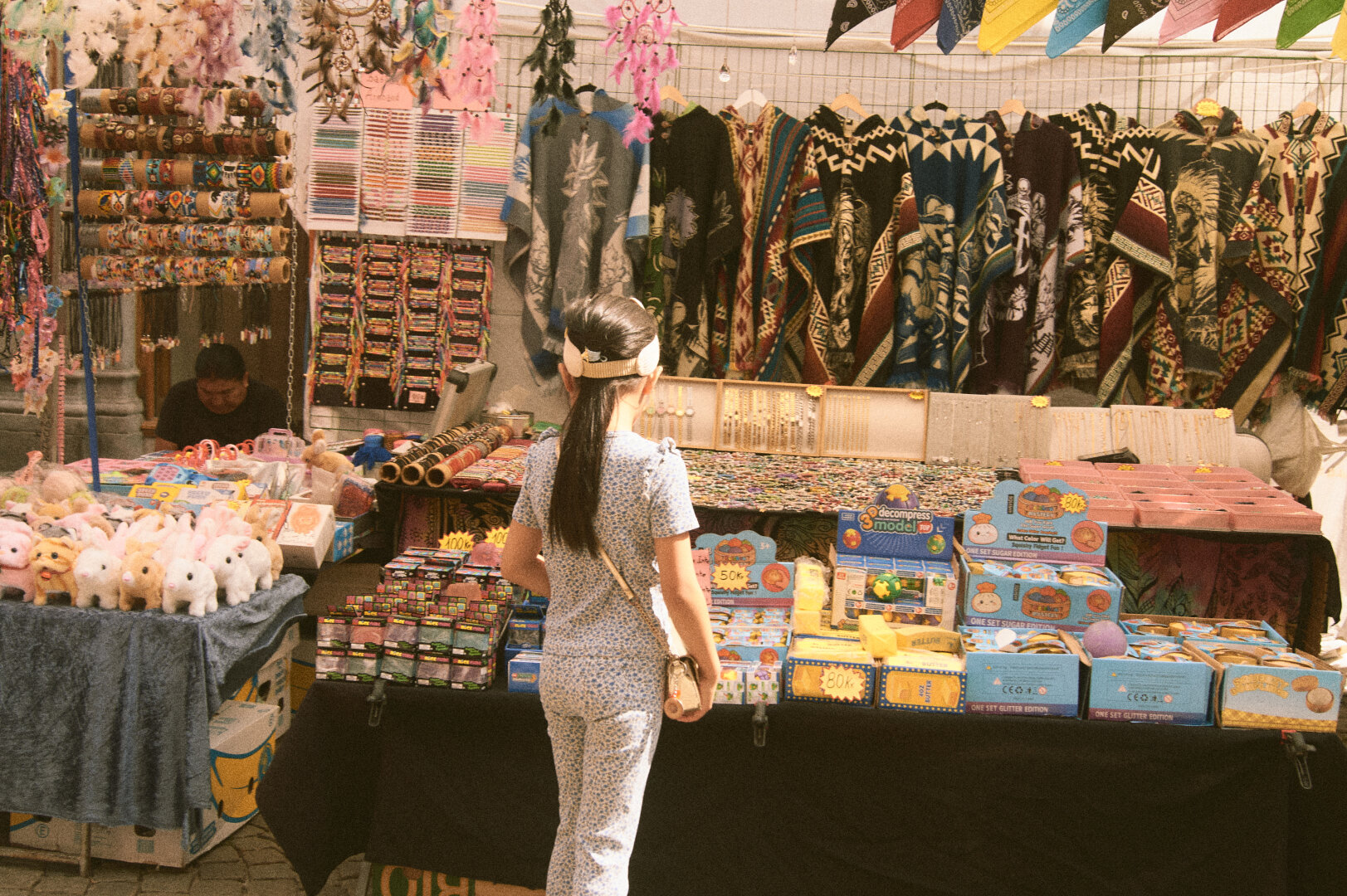 A young girl viewed from behind, looking at a vibrant outdoor market stall selling dreamcatchers, ponchos, and toys.