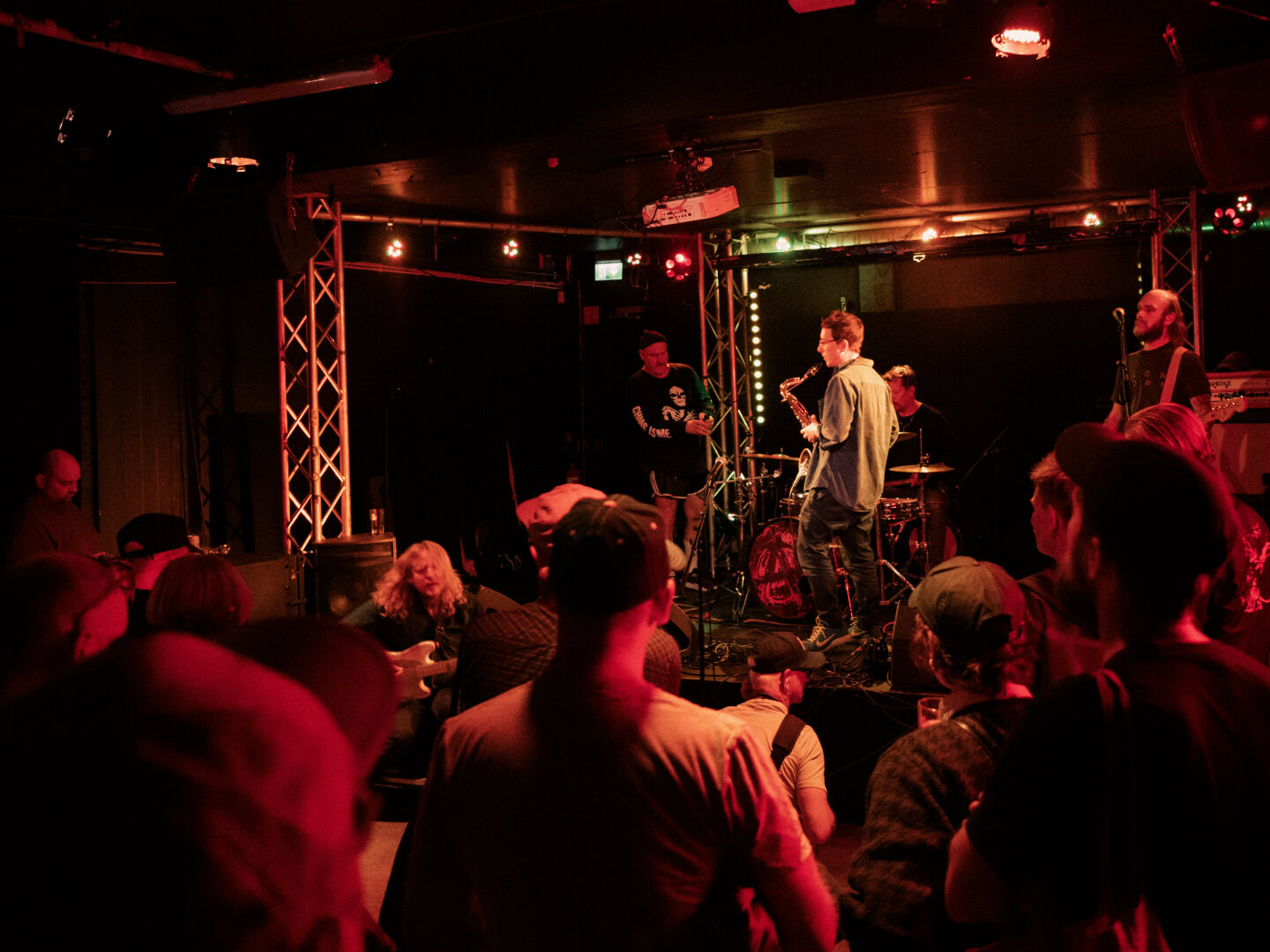 A low-light, atmospheric photograph of a live band playing on a small stage, bathed entirely in intense red lighting. The silhouettes of the audience's heads and shoulders are visible in the foreground.