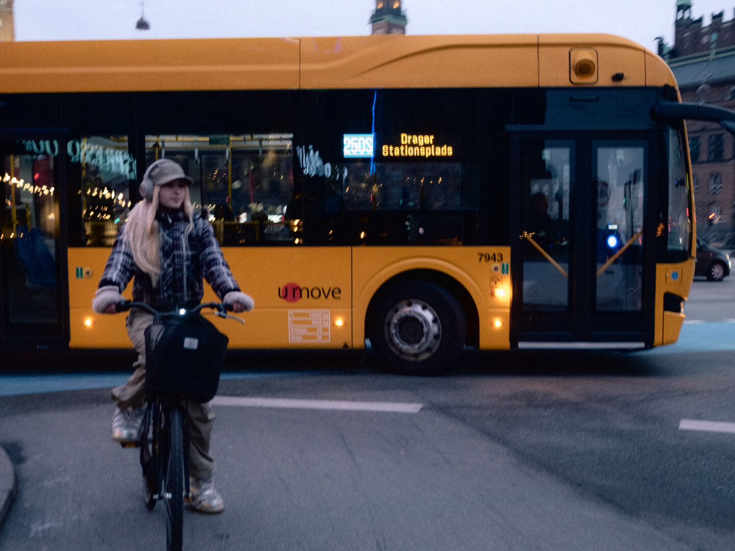 A street photograph taken at dusk in Copenhagen. A young woman wearing headphones rides a bicycle past a large yellow Movia city bus.