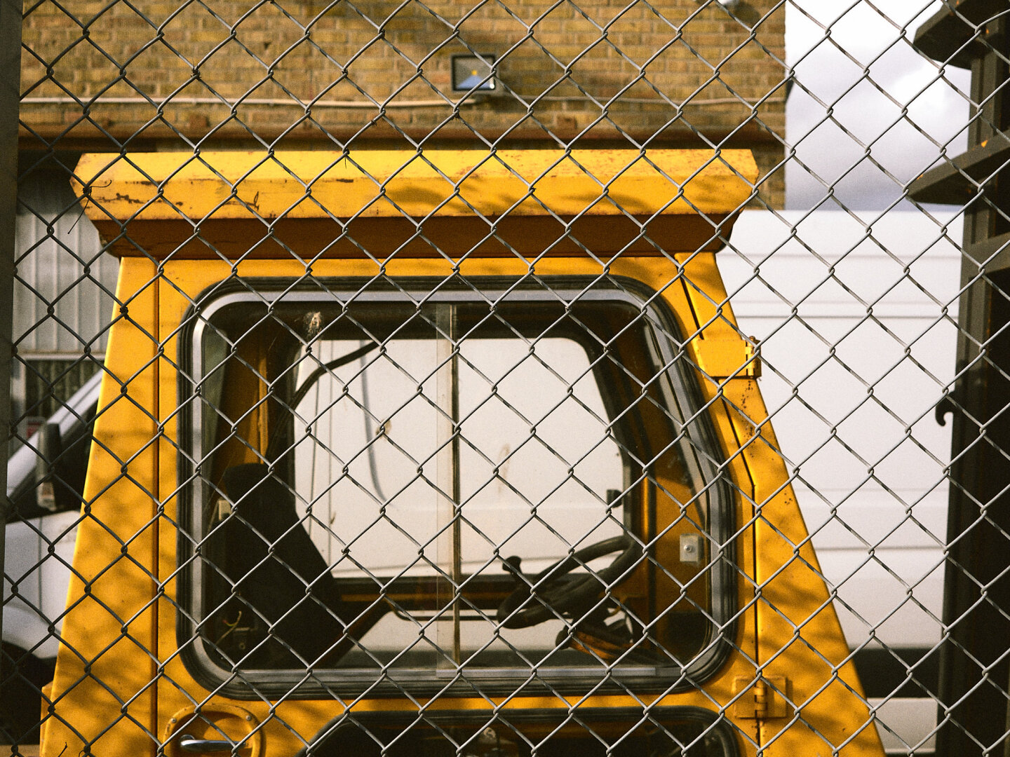 A close-up view of a bright yellow utility vehicle cab framed behind the diamond mesh of a metallic chain-link fence.