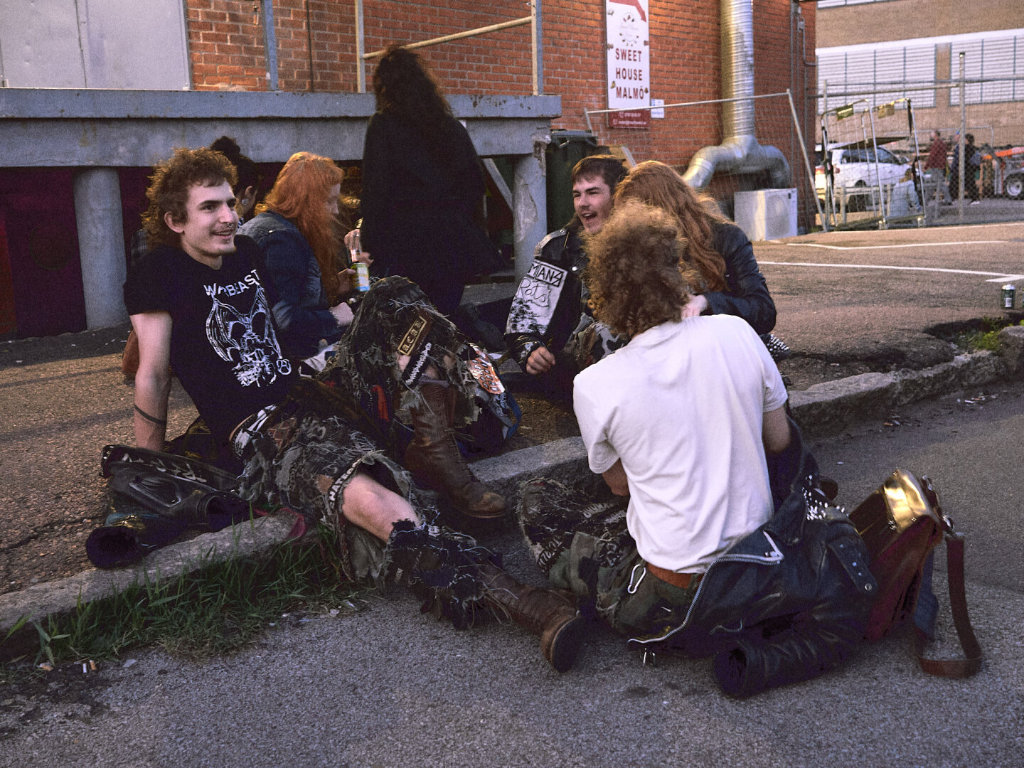 A group of alternative youth wearing patched clothes and studded leather jackets sitting on a concrete street curb, laughing and talking during the day.