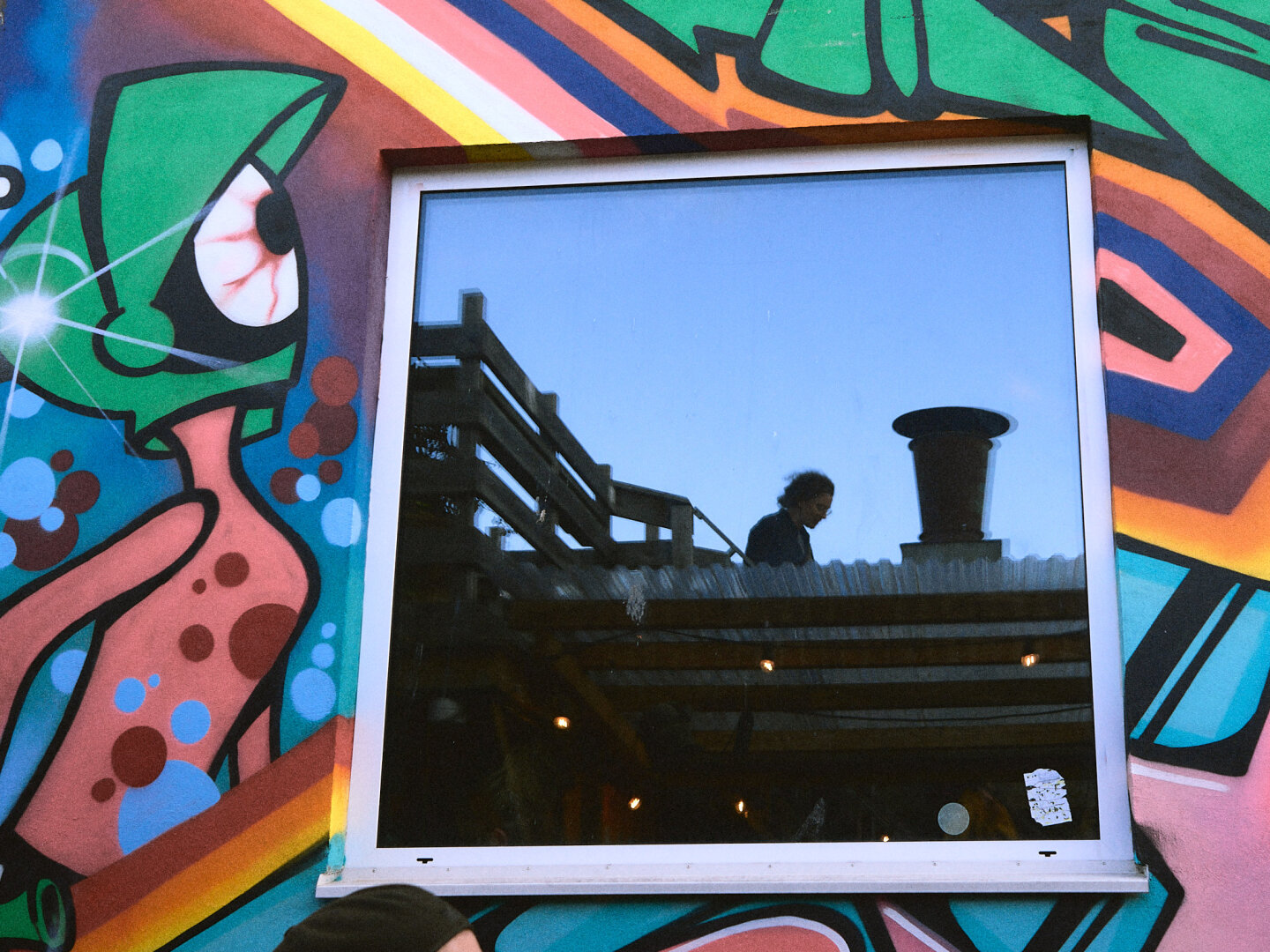A vibrant wall covered in colorful graffiti frames a square window that reflects a clear blue sky, a wooden rooftop deck, and the silhouette of a person standing beside a chimney stack.