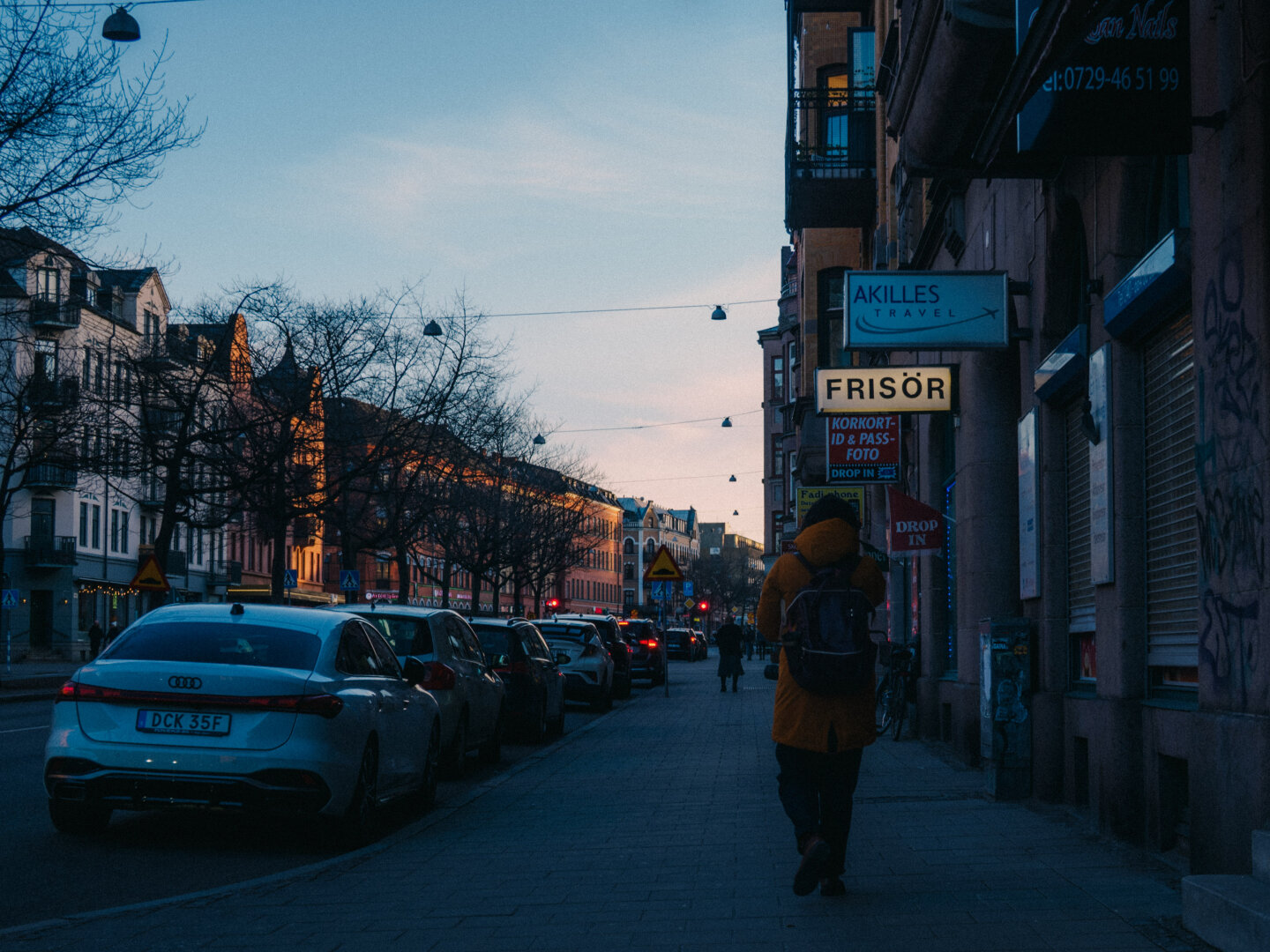 A street photography scene at dusk. A person wearing a bright yellow coat walks away down the sidewalk, flanked by parked cars on the left and shop signs on the right.