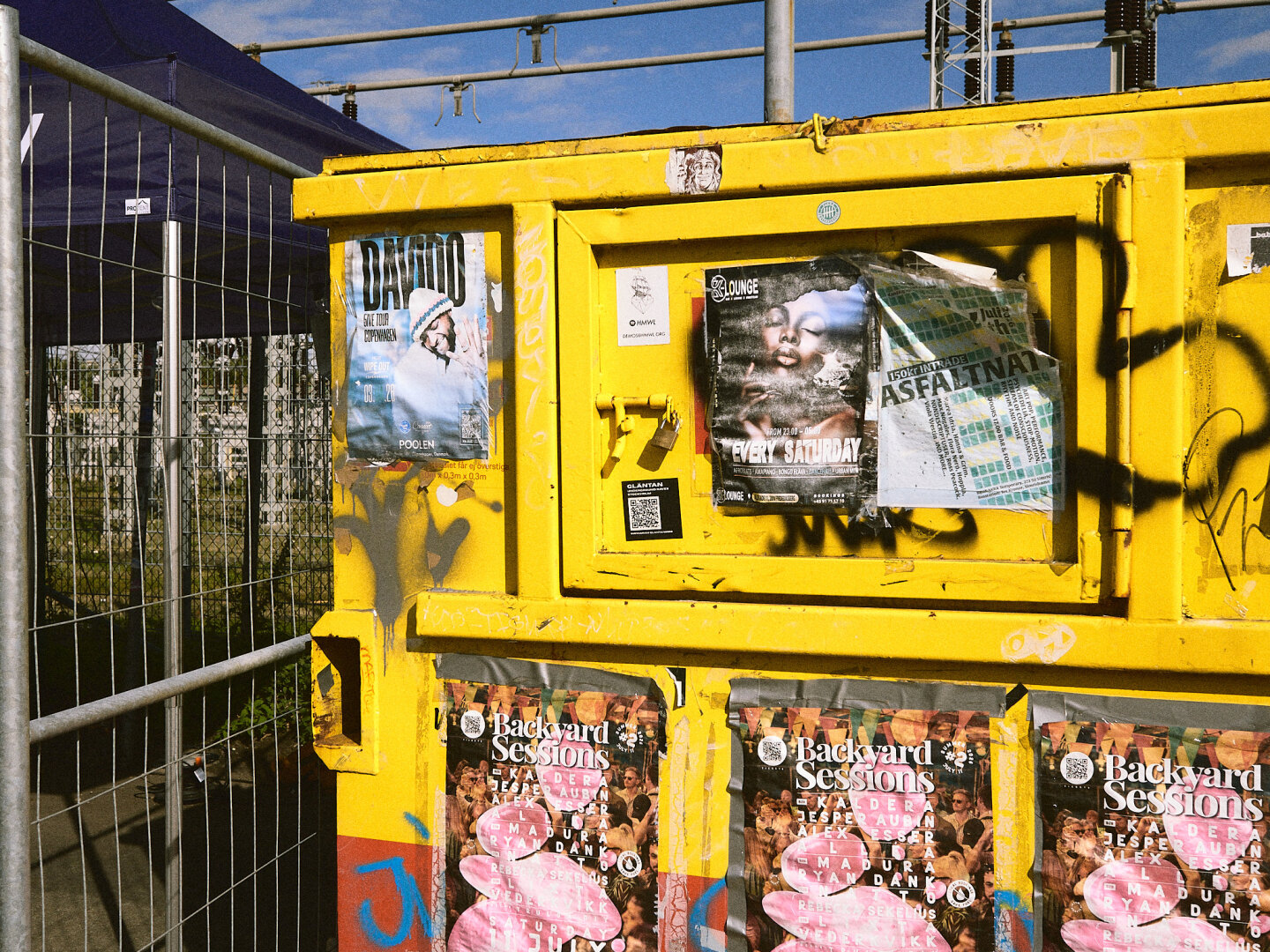 A bright yellow industrial storage box covered in layered concert posters and black spray-painted graffiti stands in direct sunlight.