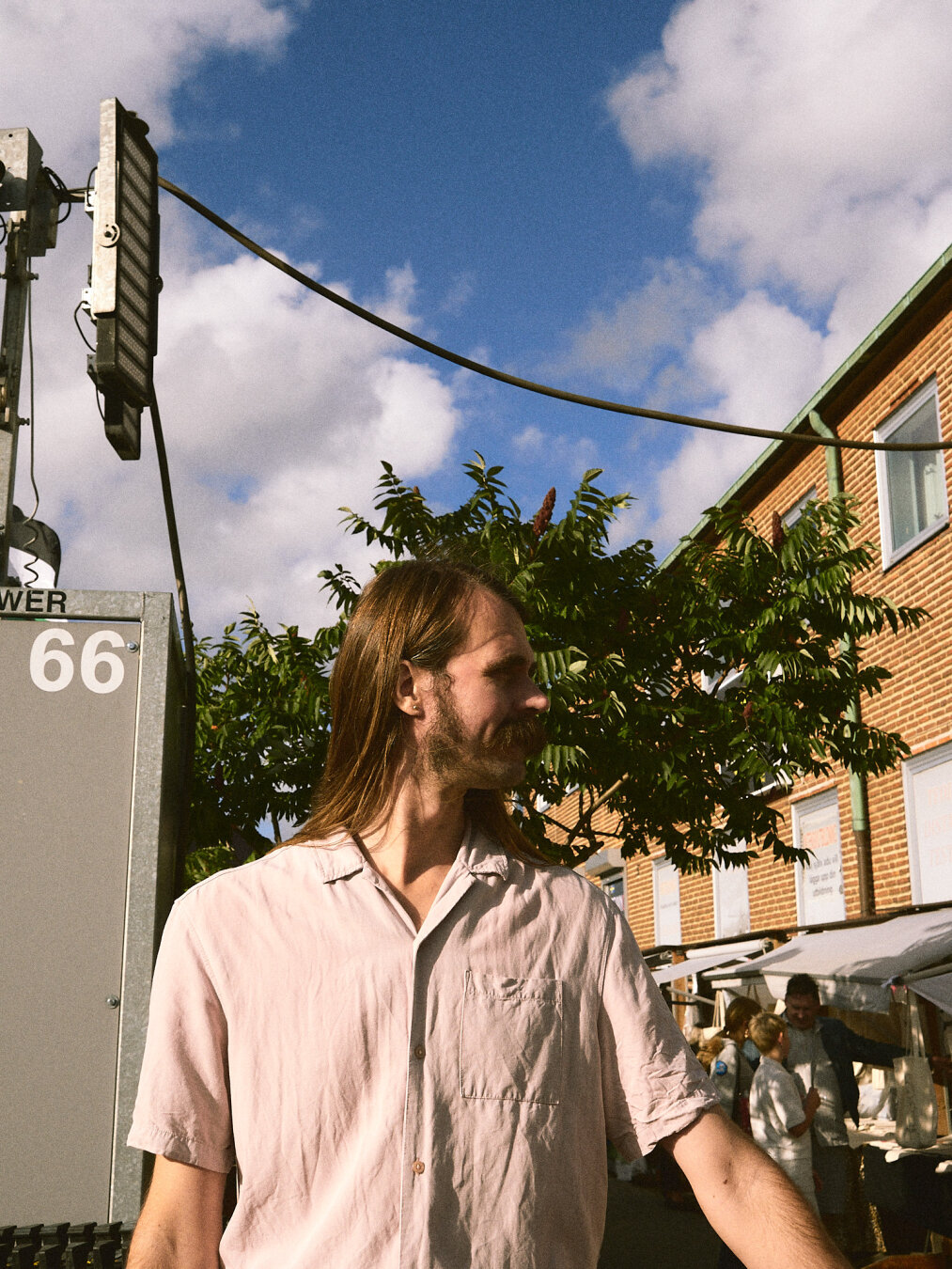 A man with long brown hair and a mustache in a light pink shirt looking to the side against a blue sky and industrial pole marked "POWER 66".