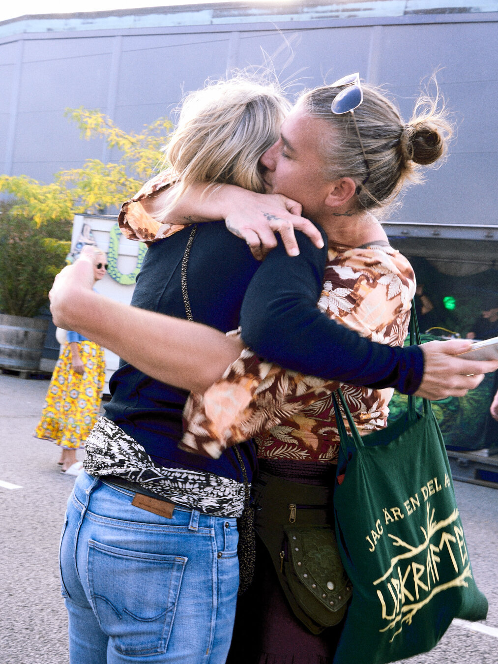 A tight, warm embrace between two people outdoors, one wearing a printed top and holding a green tote bag reading "JAG ÄR EN DEL AV UKRAFT".