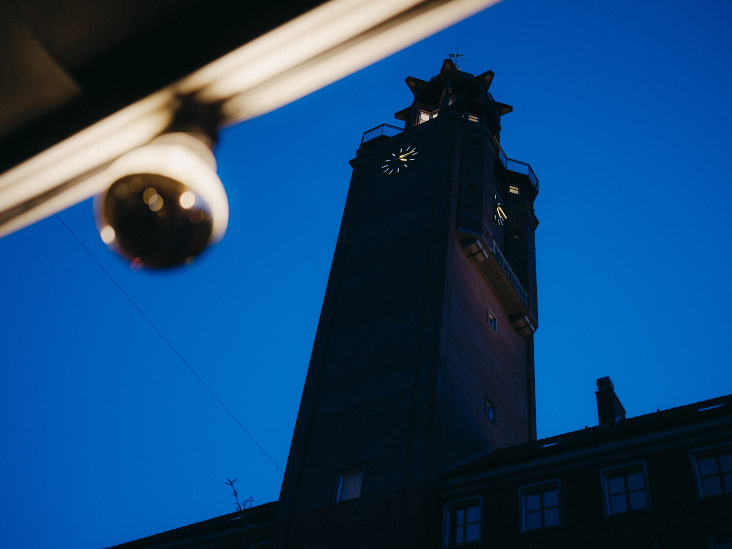 A low-angle shot of a historic brick clock tower against a deep blue evening sky. A heavily blurred modern surveillance camera looms in the foreground.