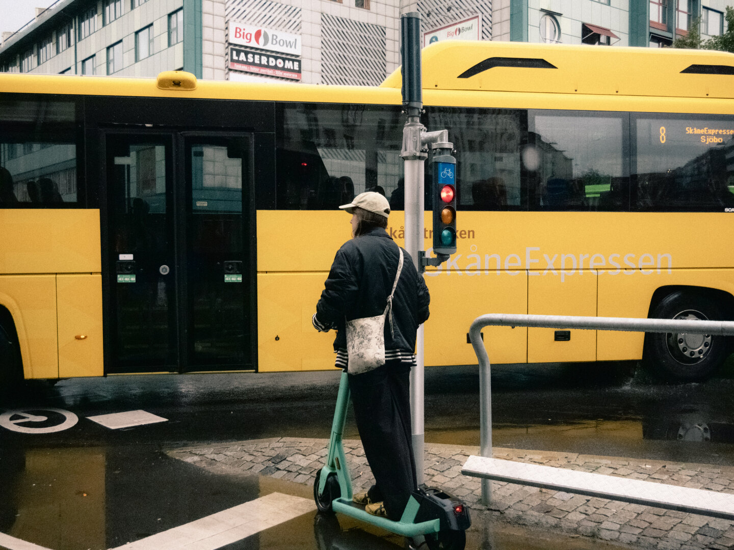 A rainy street photograph in Malmö. A person in a black jacket stands on a green electric scooter at a traffic light, framed against a bright yellow SkåneExpressen bus in the background.