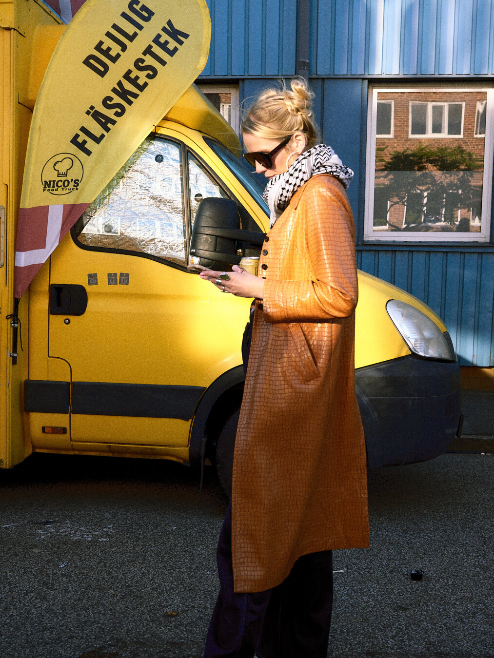 A person in a long mustard-yellow coat and keffiyeh scarf looking at a phone in front of a bright yellow food truck.