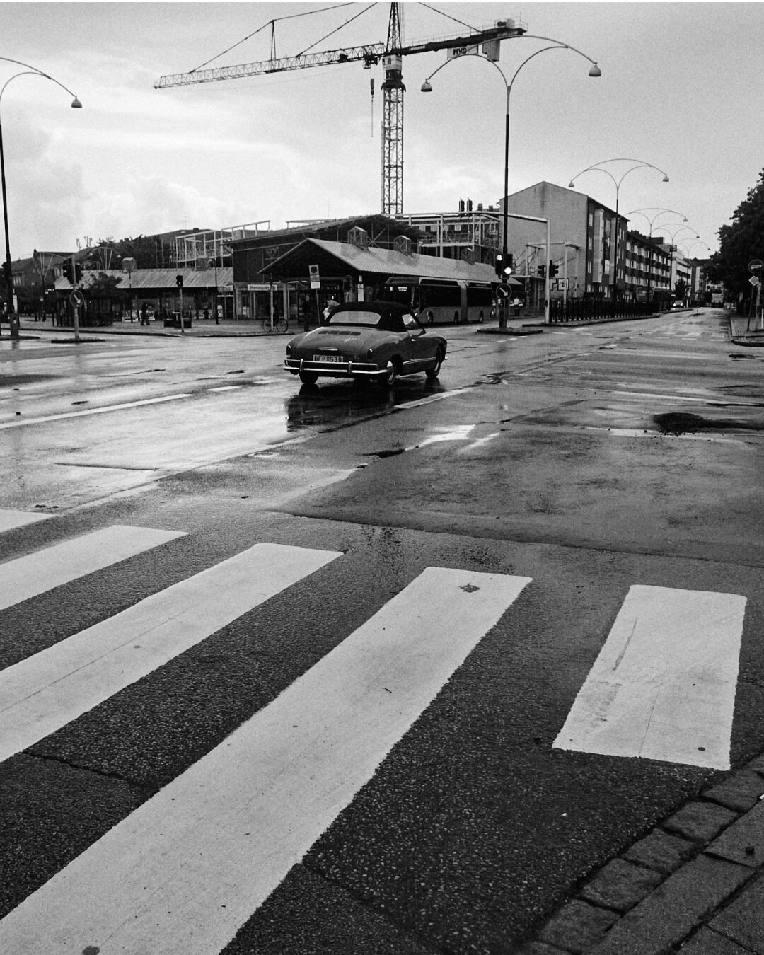 A wet city intersection with a vintage car driving away toward a construction crane under gray skies.