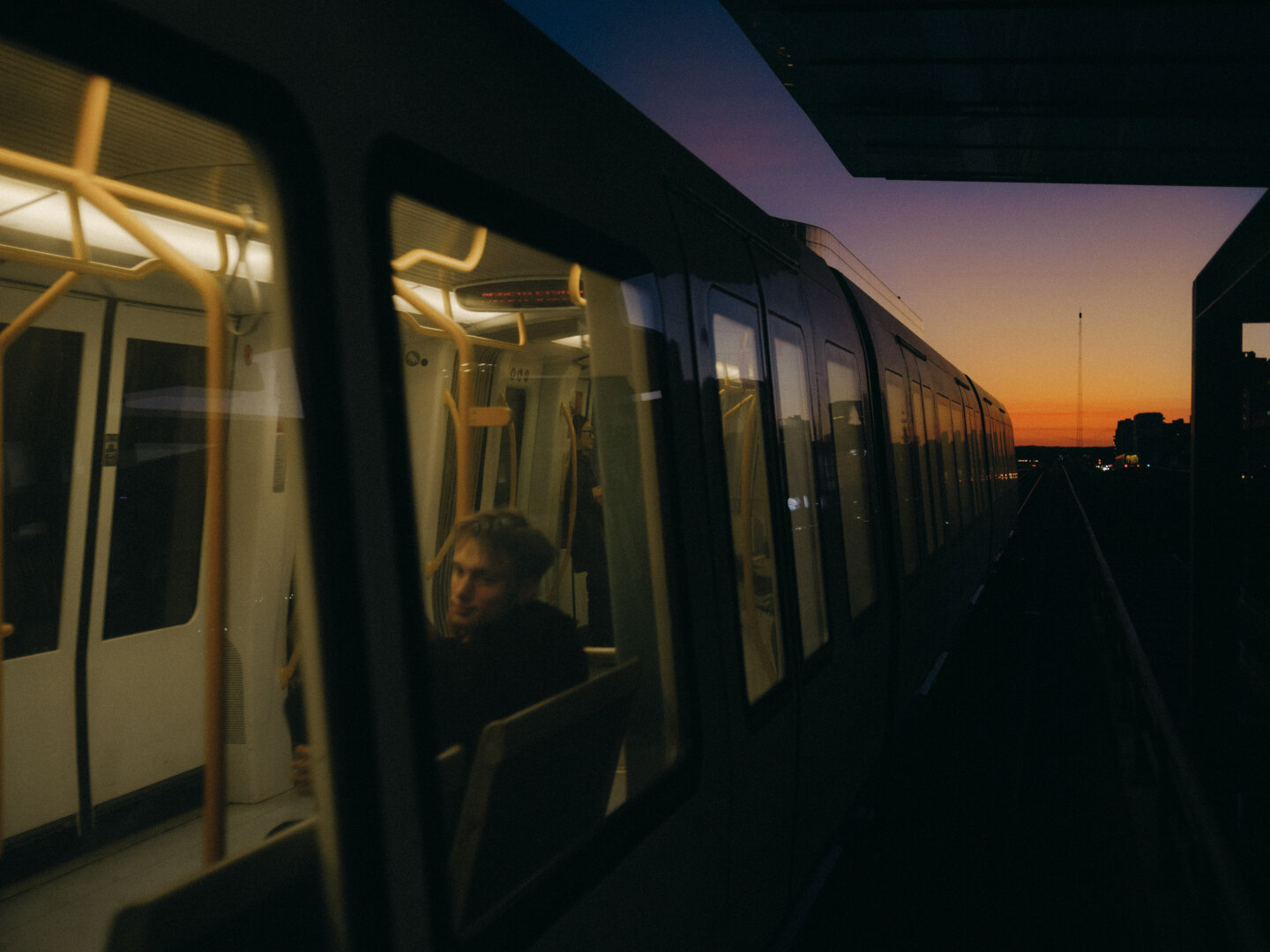 A moody twilight photograph of a metro train on a platform. Through the window, a passenger is faintly visible. The sky behind the train is a vibrant gradient of orange and purple.