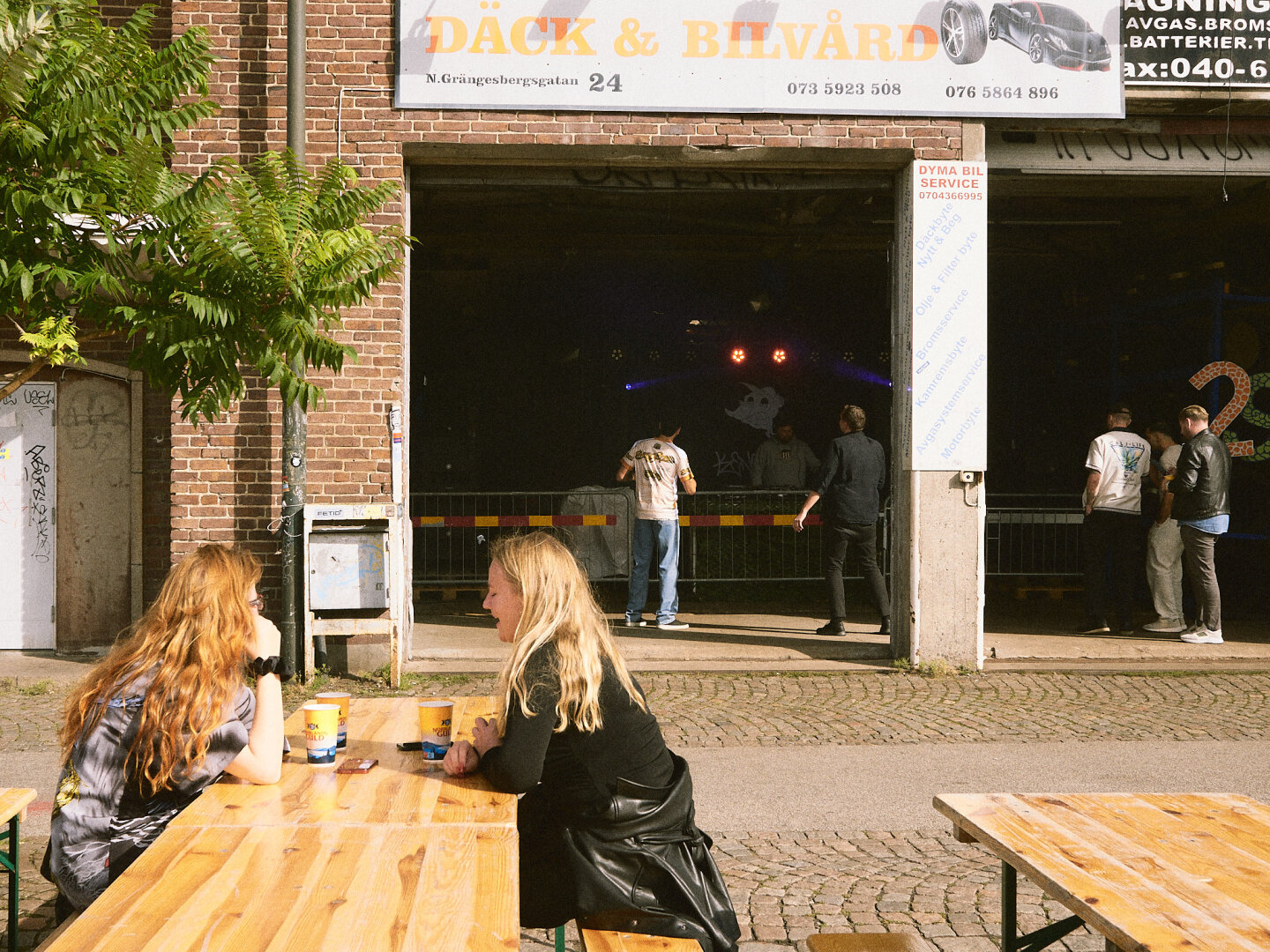 Two women sit at a wooden picnic table outside an industrial brick building with a sign that reads "Däck & Bilvård", where a DJ performs inside the open garage bay under stage lights.
